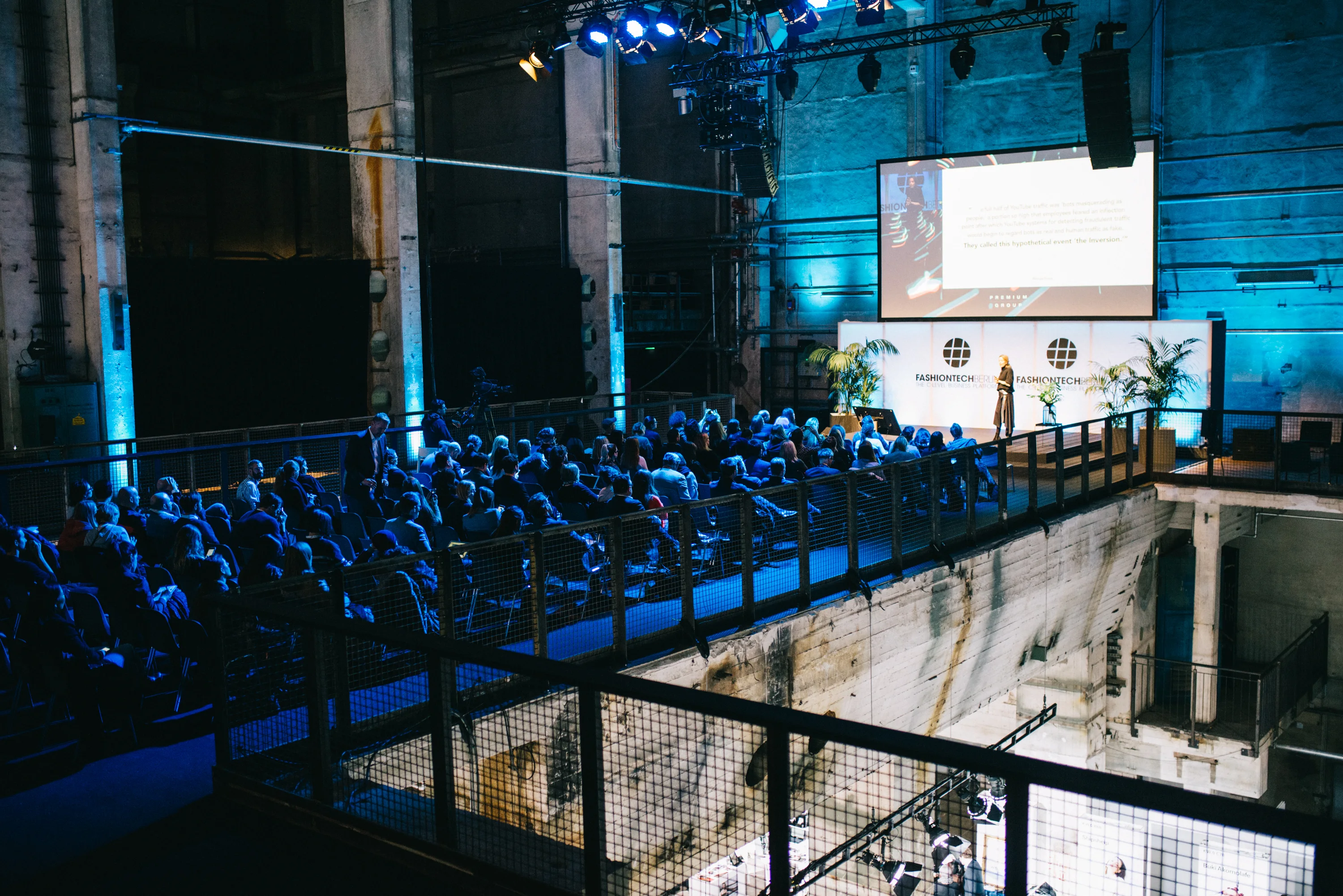 Audience on a gallery level watching a FASHIONTECH Berlin keynote in blue light, with the speaker beside a large screen.