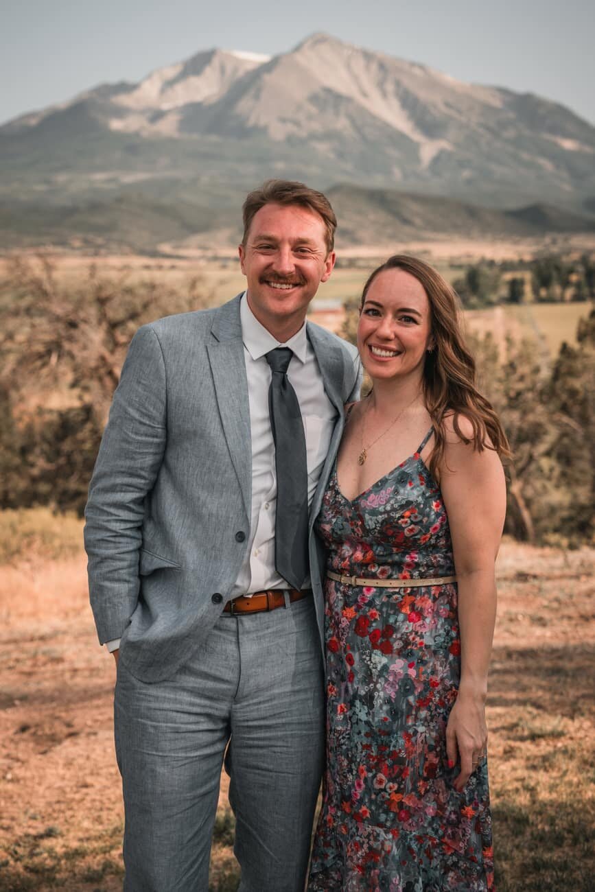 Couple in a gray suit and floral dress posing in front of a mountain peak