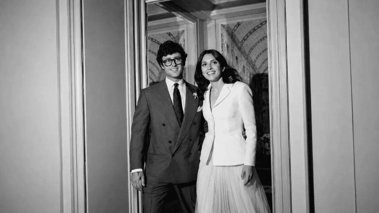 A black-and-white photo of a couple posing together in formal attire, smiling in a doorway.
