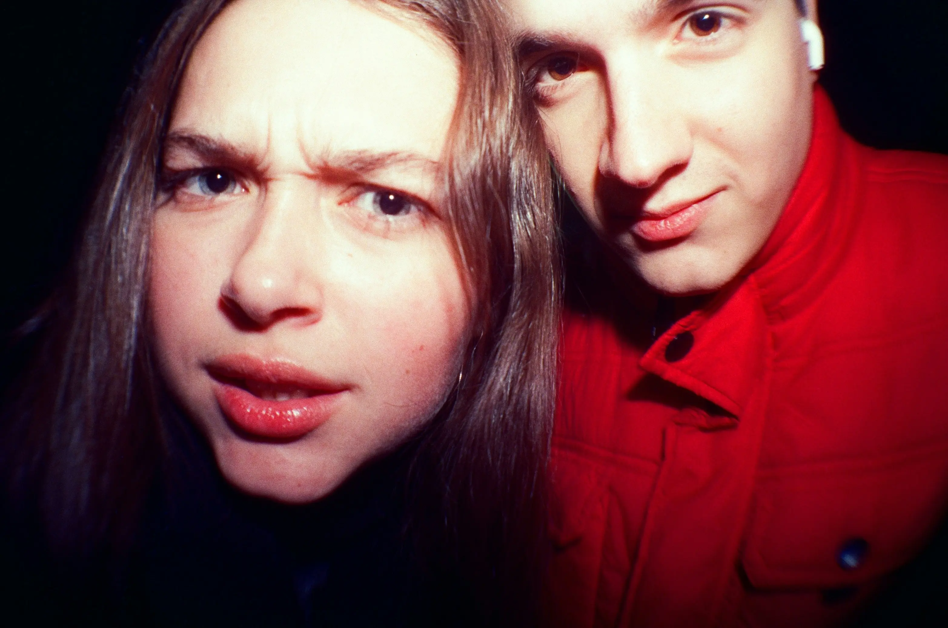 Candid close-up portrait of two young adults looking directly at the camera with intense expressions, set against a black background.