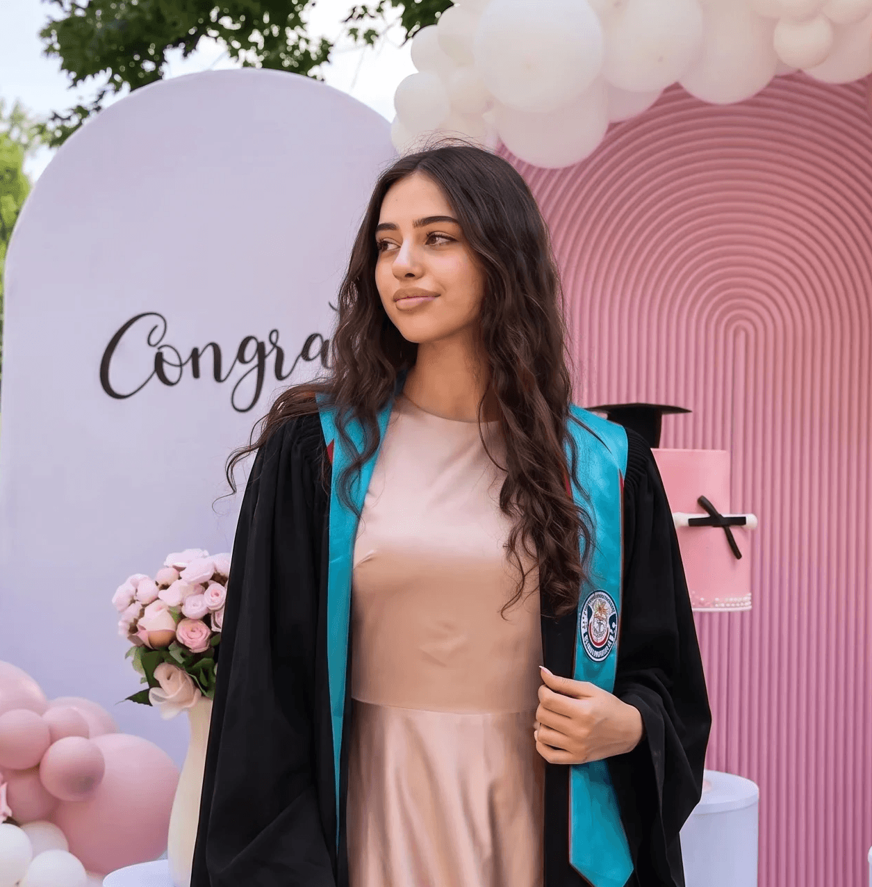 Young woman in a graduation gown beside a pink and white balloon arch Graduation party setup with balloons, roses, and a Congratulations backdrop