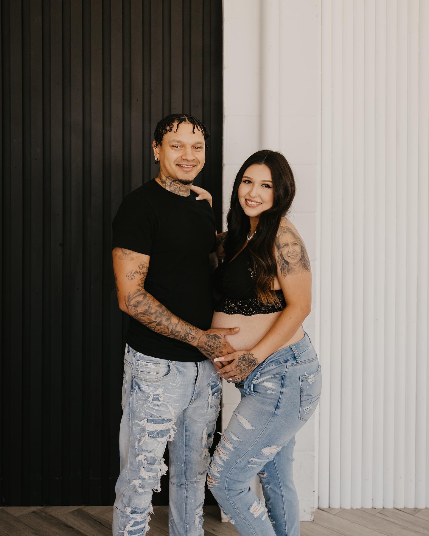 A couple poses affectionately against a contrasting black and white corrugated wall. The man, adorned with tattoos and wearing a black t-shirt, gently embraces the woman, who smiles in a black lace top and distressed jeans, hinting at a casual yet intimate moment between them.