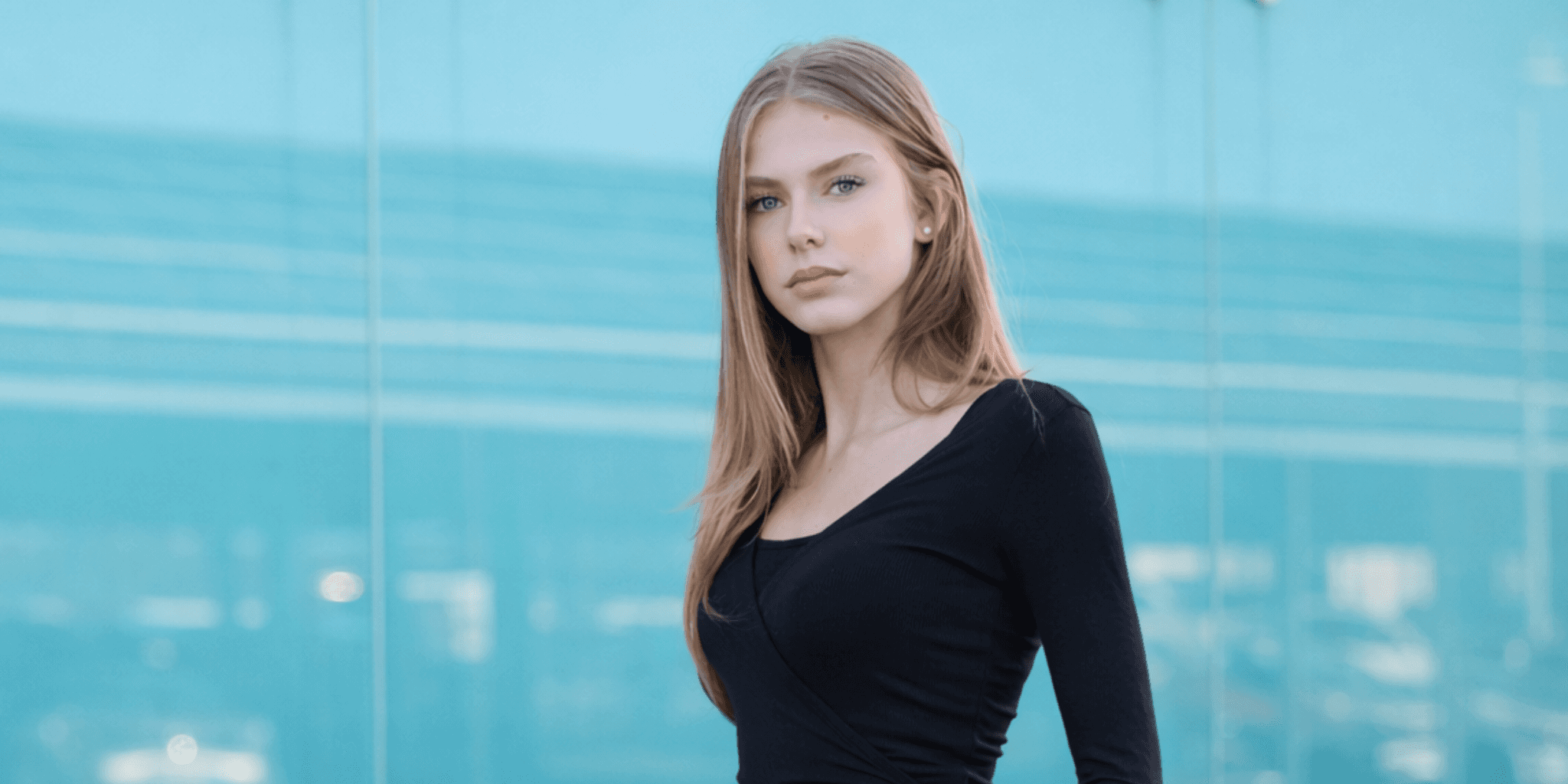 Young woman with long hair in a black top stands against a glass wall backdrop. The soft, blue tint and her calm expression create a serene mood.