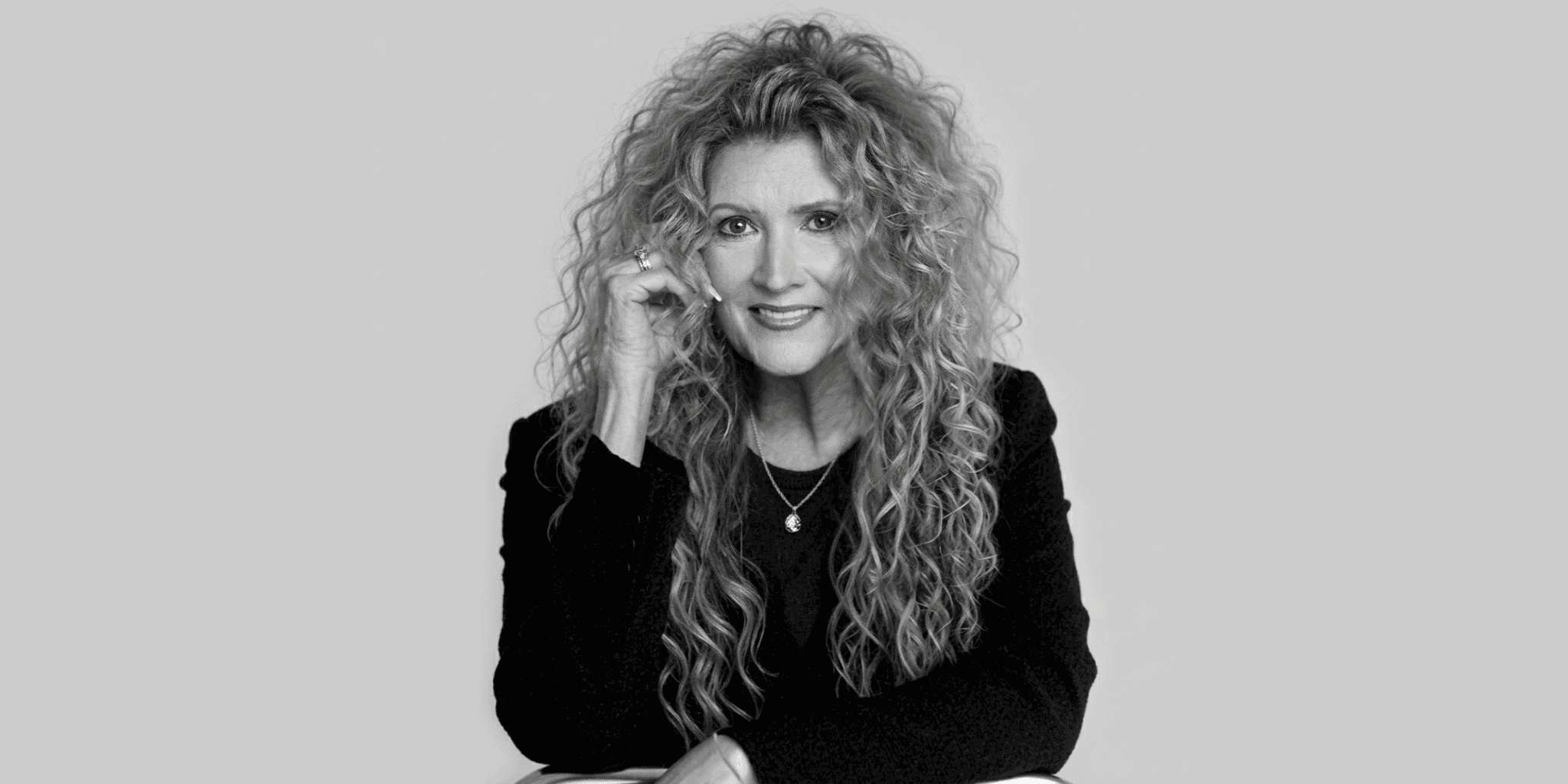 A woman with long, curly hair smiles softly, resting her head on her hand. She's wearing a black top and necklace, set against a light gray backdrop.