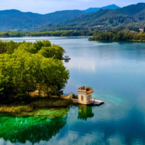 A small, yellow building on a pier by a clear, tranquil lake surrounded by lush greenery and distant mountains.