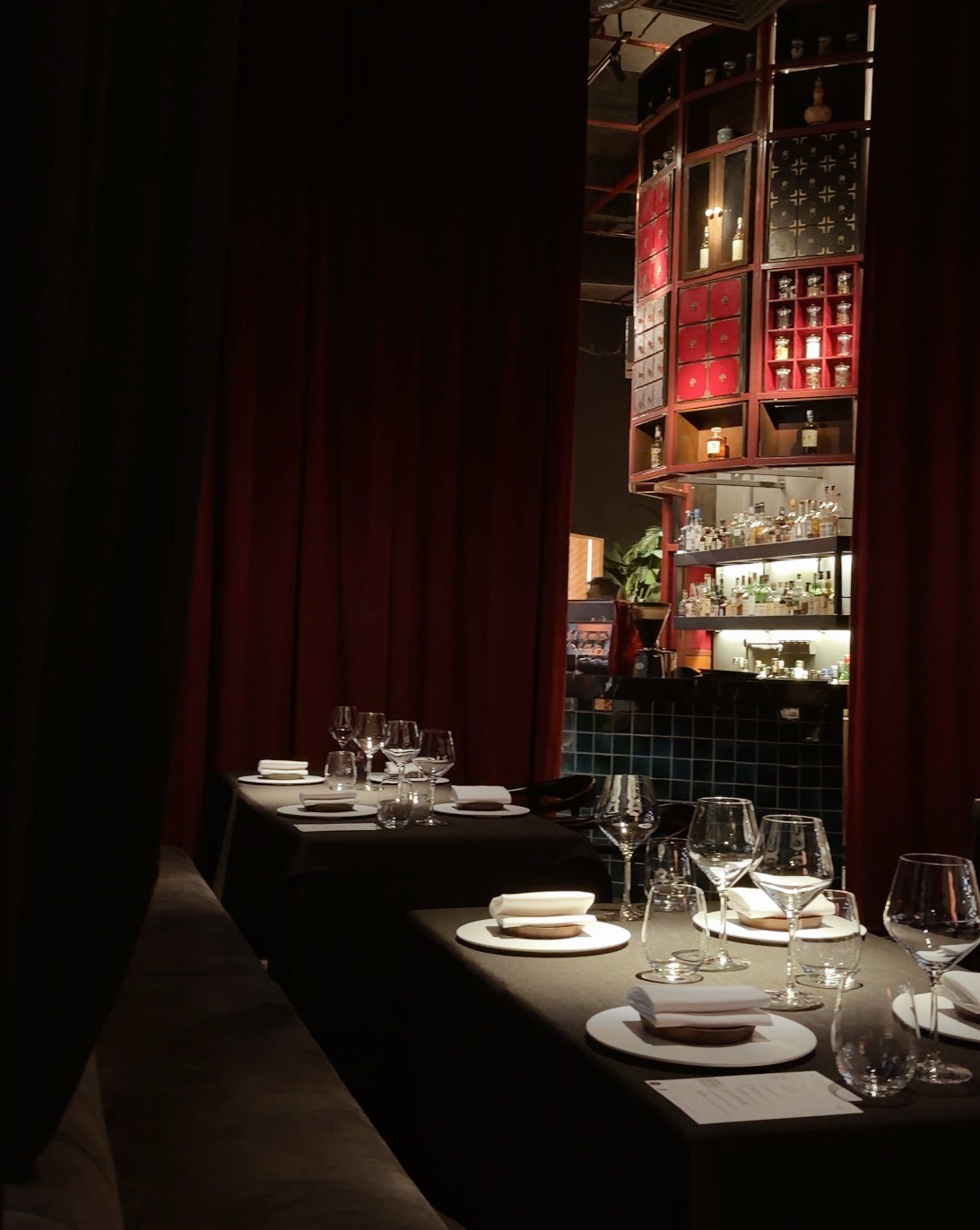 A dimly lit dining room at BETA KL featuring elegantly set tables with glassware and plates, framed by rich red curtains and a well-stocked bar in the background.