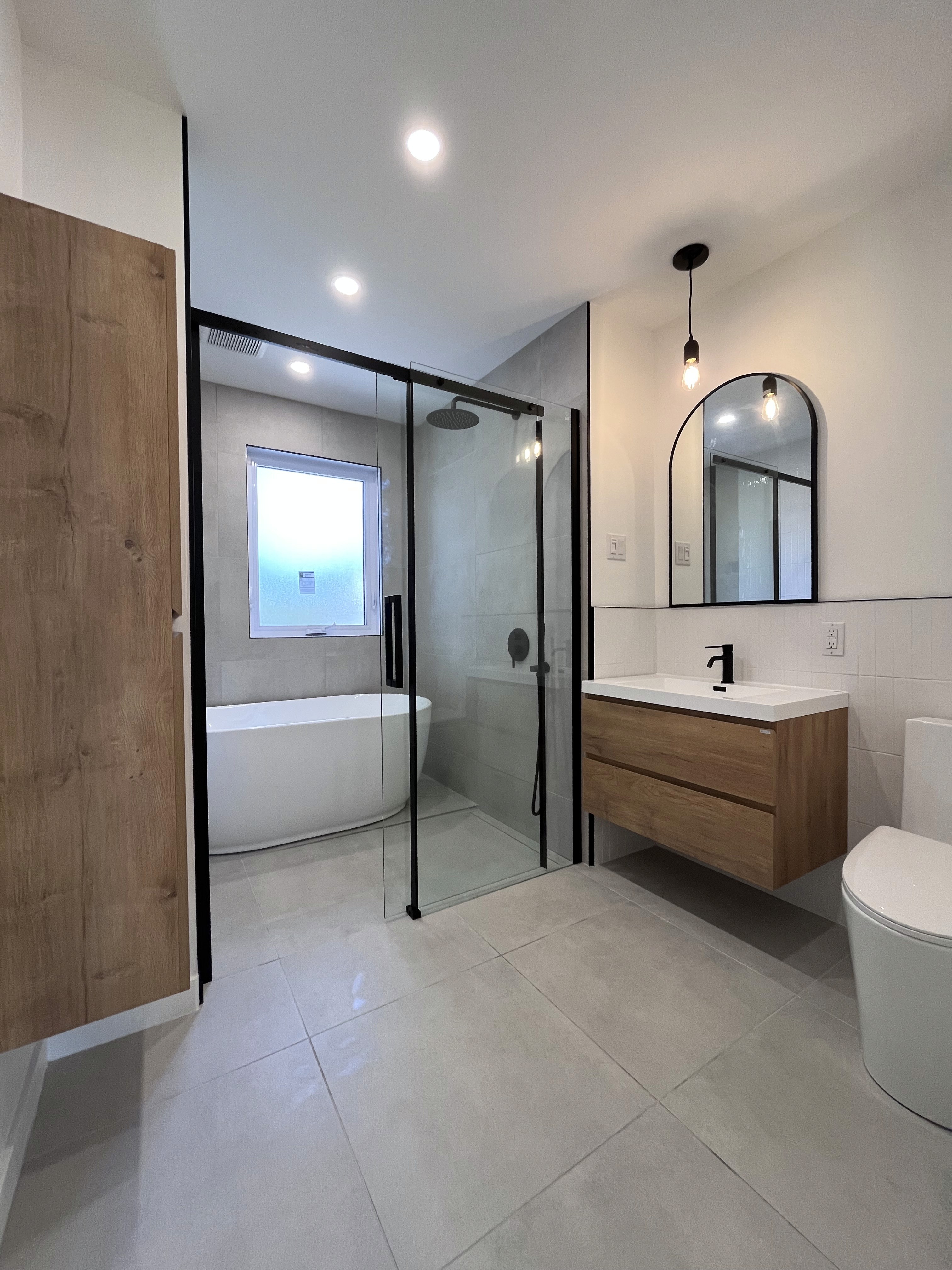 Salle de bains aux teintes de gris et de crème. La céramique blanche lustrée aux murs met en valeur la porcelaine grise et beige au sol dans la douche. La vanité et l,armoire penderie en bois sont accompagnées d'un miroir en forme d'arche. Le bain et la douche sont réunis afin d'optimiser l,espace et d'offrir une excpérience à la fois fonctionnelle et enveloppante.