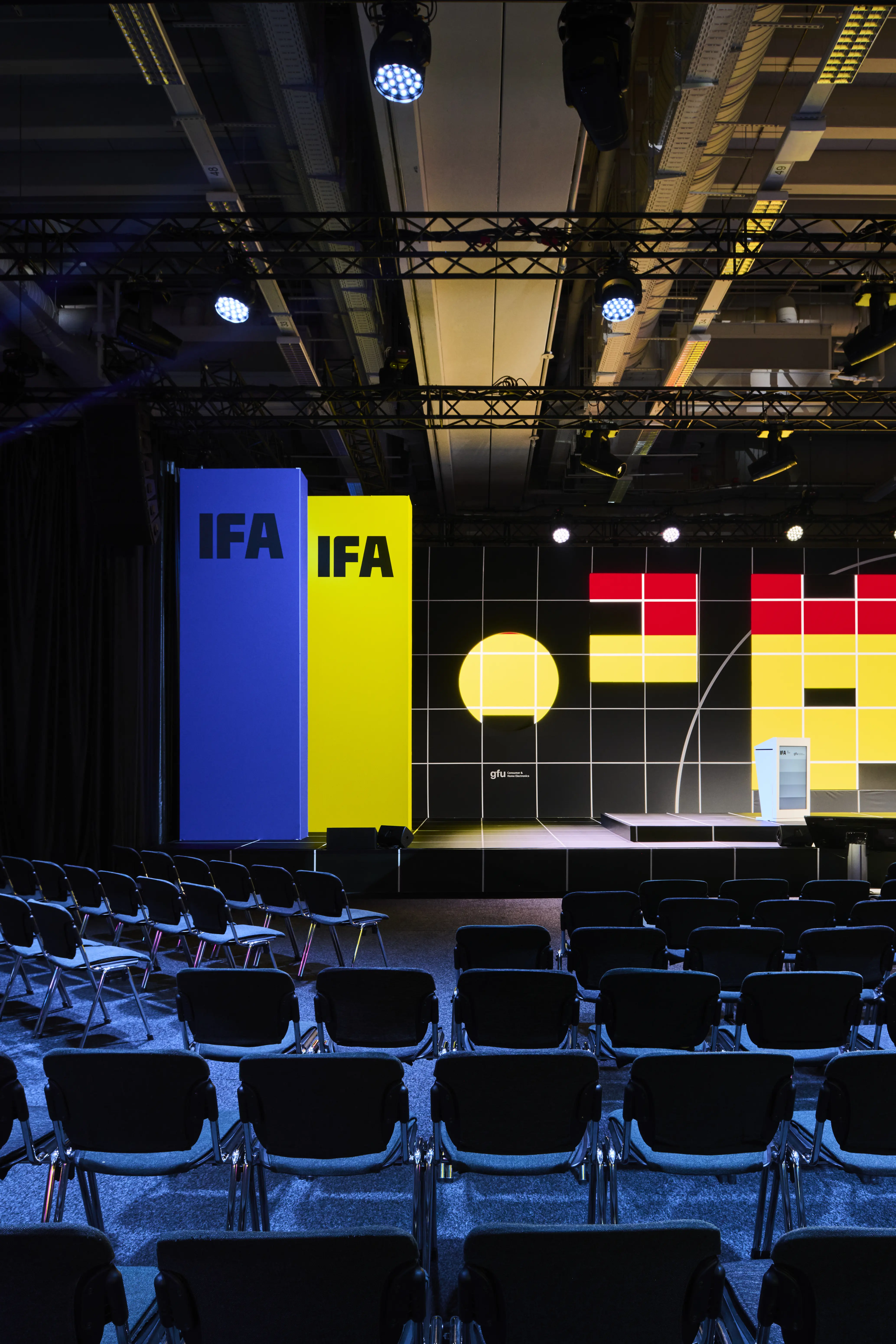 Blue and yellow IFA pillars in front of the stage with graphic LED wall and seating.