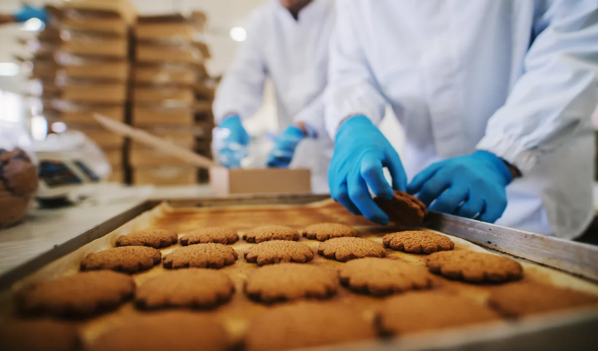 Gloved hands lifting a biscuit from a baking tray while staff pack in the background