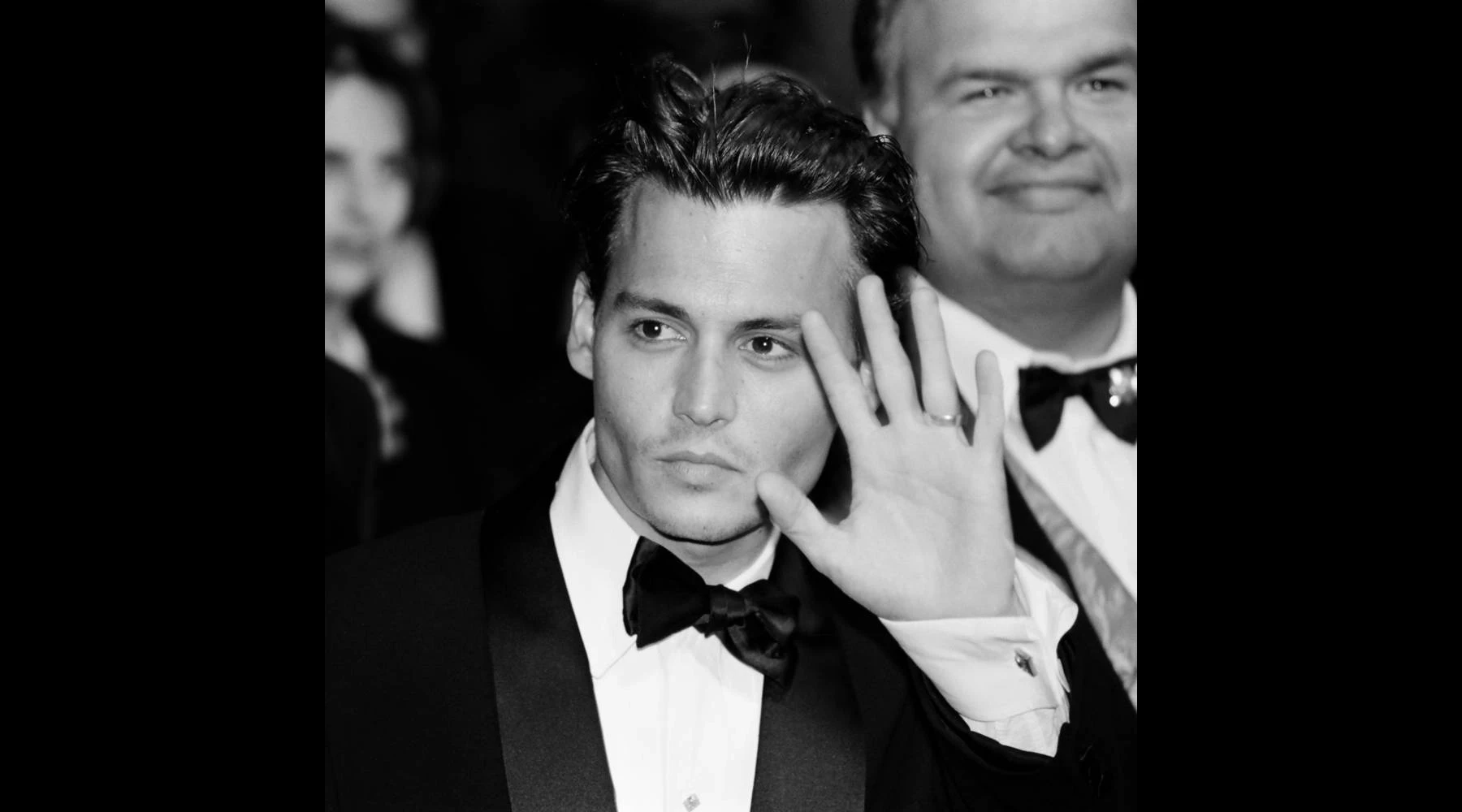 A man in a tuxedo raising one hand toward the camera, his face partially obscured behind his palm, other figures visible and out of focus behind him, black and white