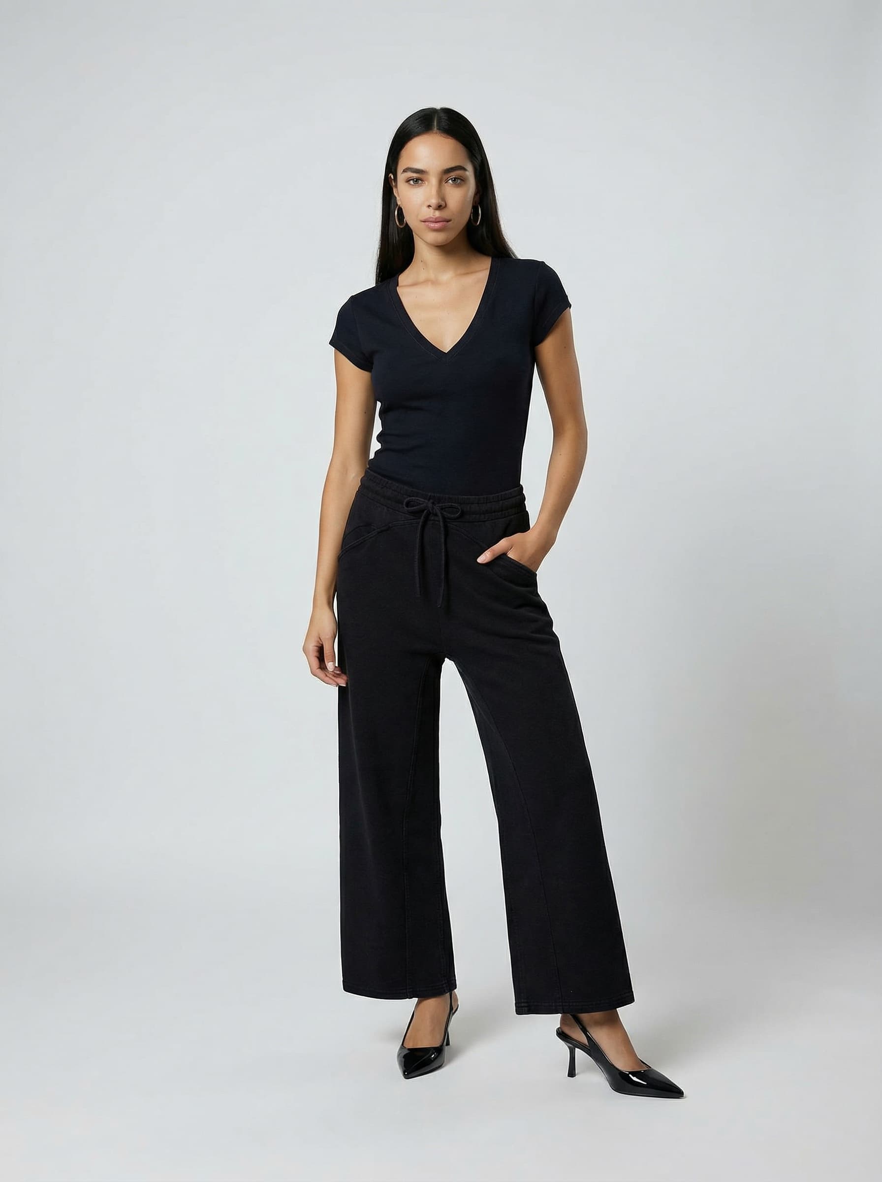 A woman standing against a plain backdrop, wearing a black short-sleeved top and wide-leg trousers, paired with black heels, exuding a minimalist and stylish fashion look.