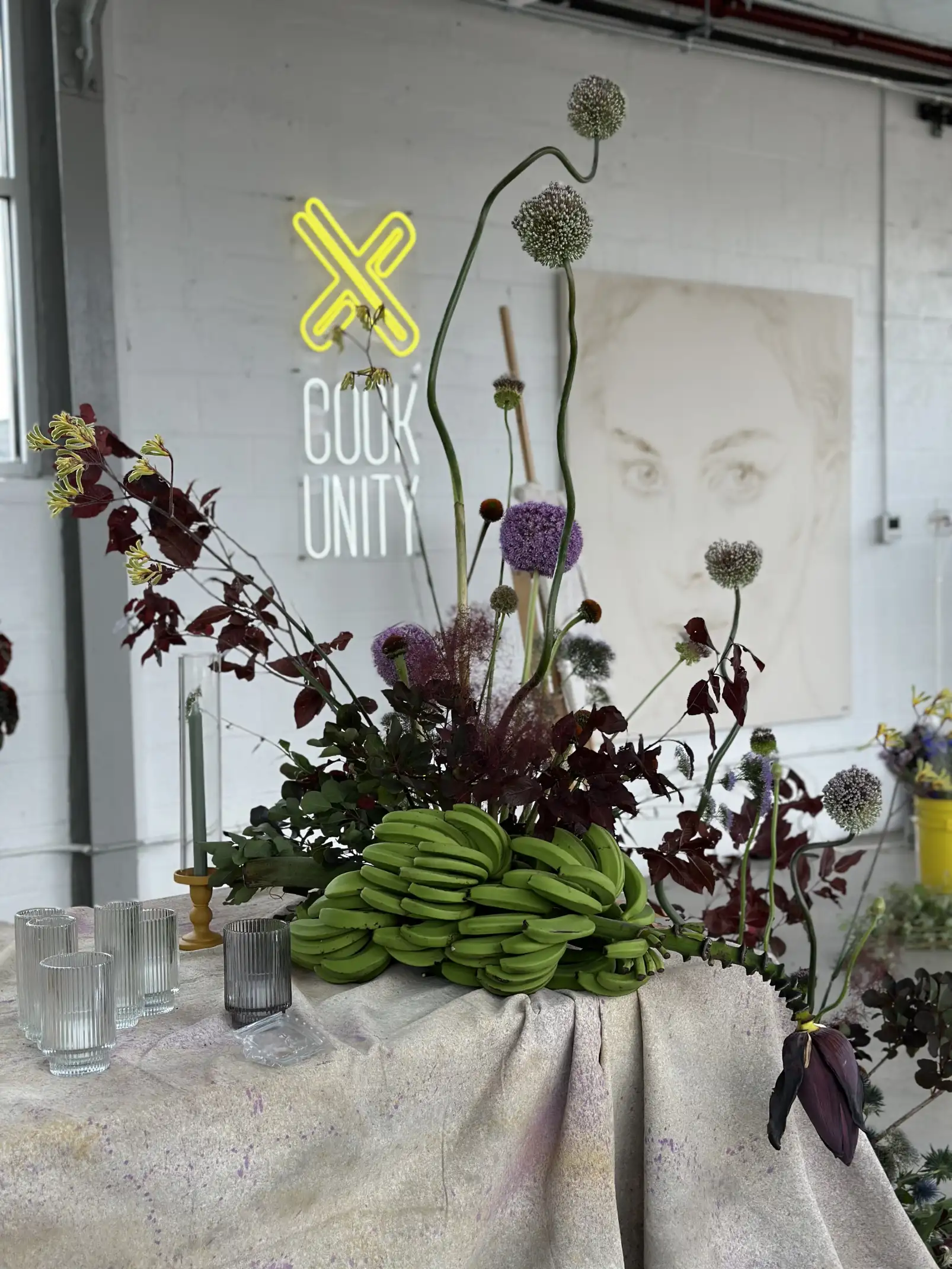 Arreglo decorativo con racimos de bananas verdes, flores altas en tonos morado y blanco y follaje oscuro sobre una mesa con mantel, en un espacio con el logo luminoso de Cook Unity al fondo.