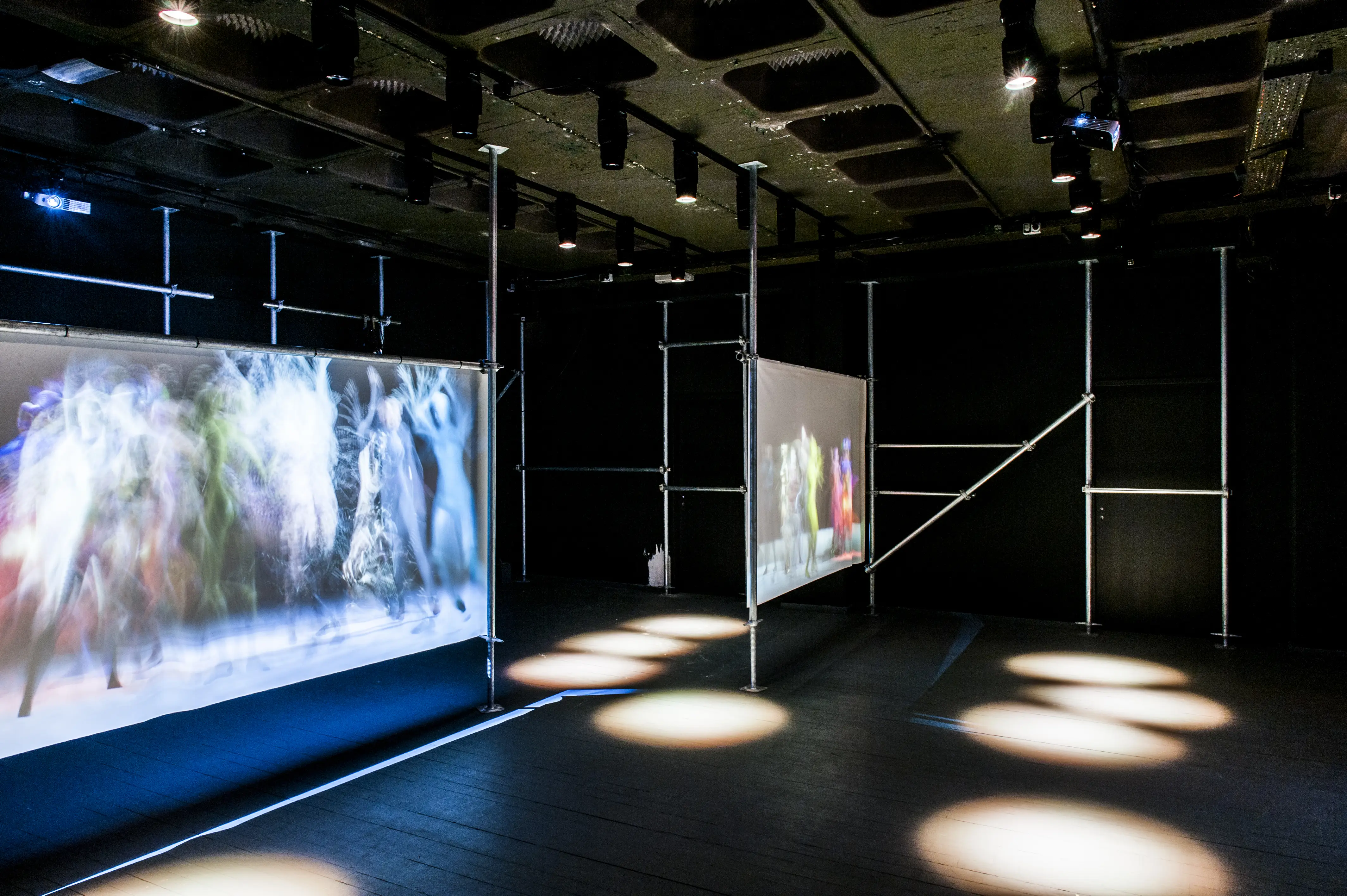 Dark event space with two scaffold-mounted projection screens showing dancers and round light spots on the floor.