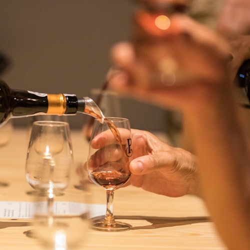Mãos servem e seguram taças de vinho rosé em um evento de degustação, com taças adicionais vazias e outras pela metade sobre uma mesa de madeira clara.