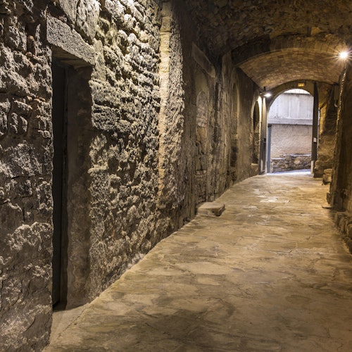 一条狭窄、光线昏暗的石头通道,拥有拱形天花板和磨损的石墙,通向一个开阔、光线较好的区域。