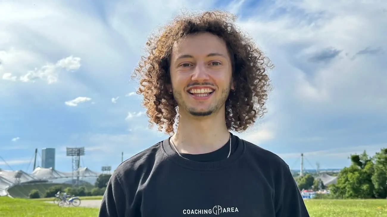 Thiago Calderaro, Founder and CEO of CoachingArea, smiles against the backdrop of a sunny park with stadium structures in the distance. Wearing a black CoachingArea sweatshirt, Thiago embodies the spirit of innovation and community in amateur sports. This image represents the welcoming and inclusive vision of CoachingArea, aiming to connect sports enthusiasts and improve the organisation and development of amateur sports through a comprehensive digital platform.