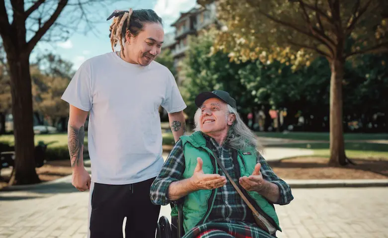 A younger man and an older adult in a wheelchair sharing a lighthearted, laughing moment together outdoors on a sunny day.
