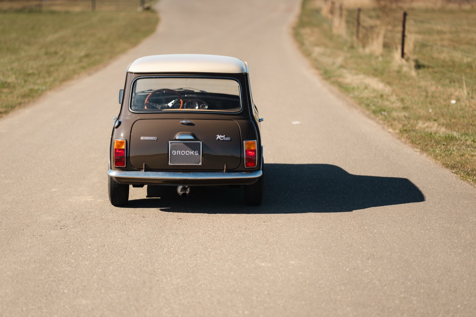 A vintage mini classic car drives down a winding road, casting a shadow on the pavement under clear skies.