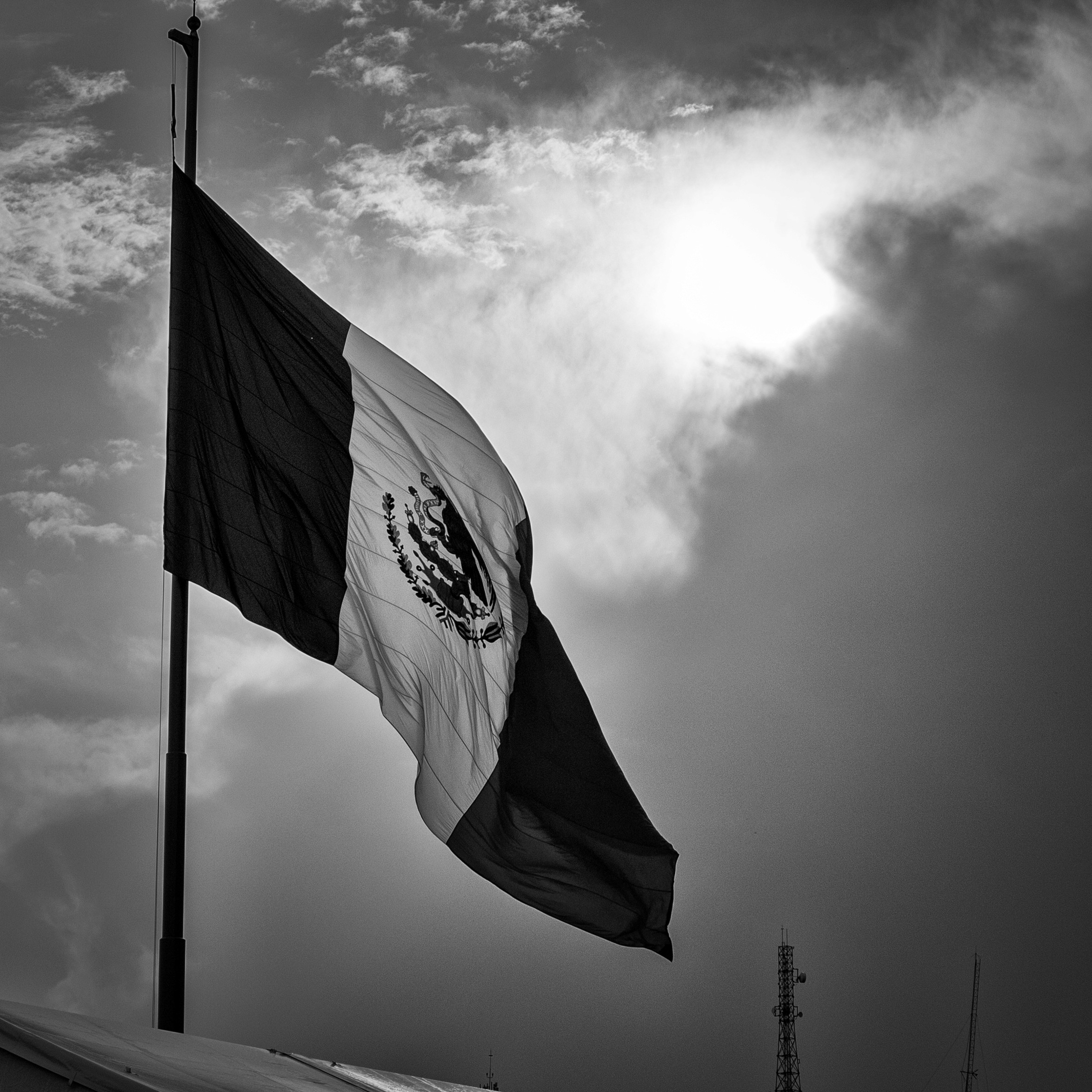 Black and white photo of the Mexican flag billowing in the wind against a moody sky with sun breaking through the clouds.
