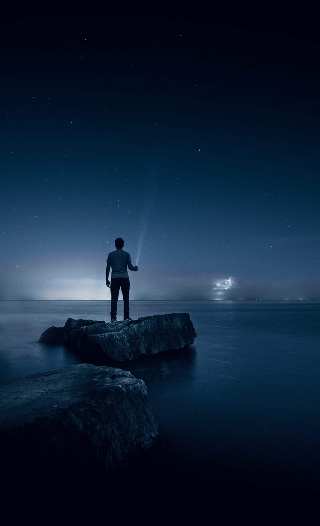 A man shining a light into the sky standing on rocks at the edge of a lake