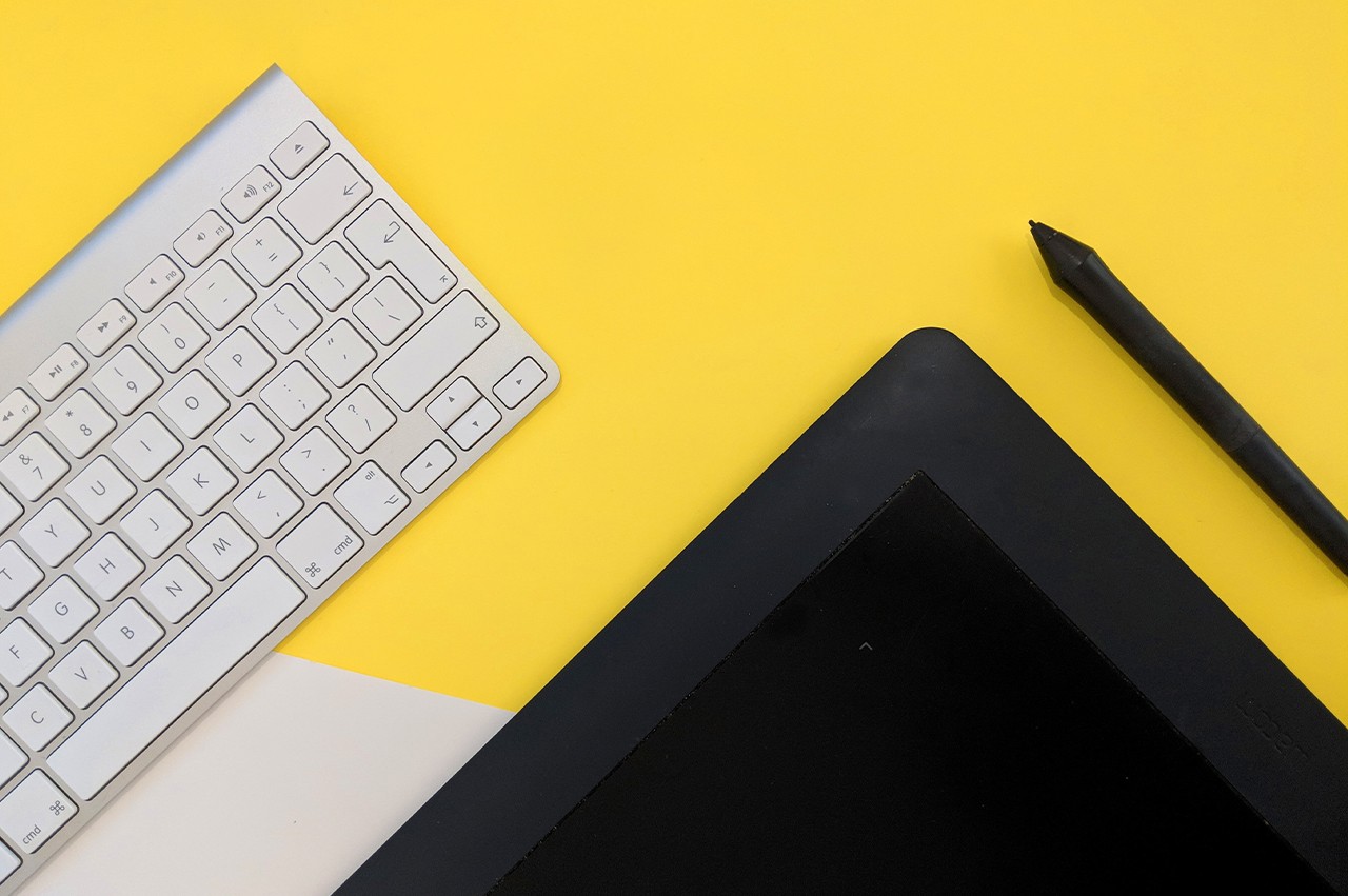 Overhead view of a yellow desk with an Apple keyboard and graphic tablet with pen