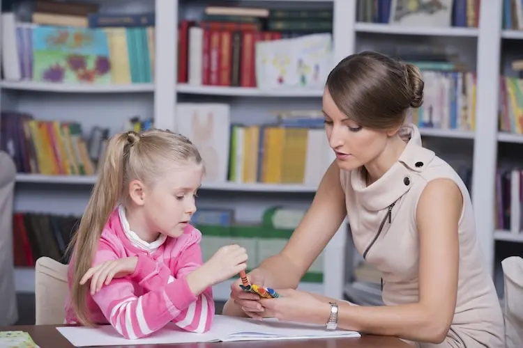 Female teacher helps young girl with colorful blocks during a learning activity in a classroom filled with books, promoting early childhood education and tutoring.