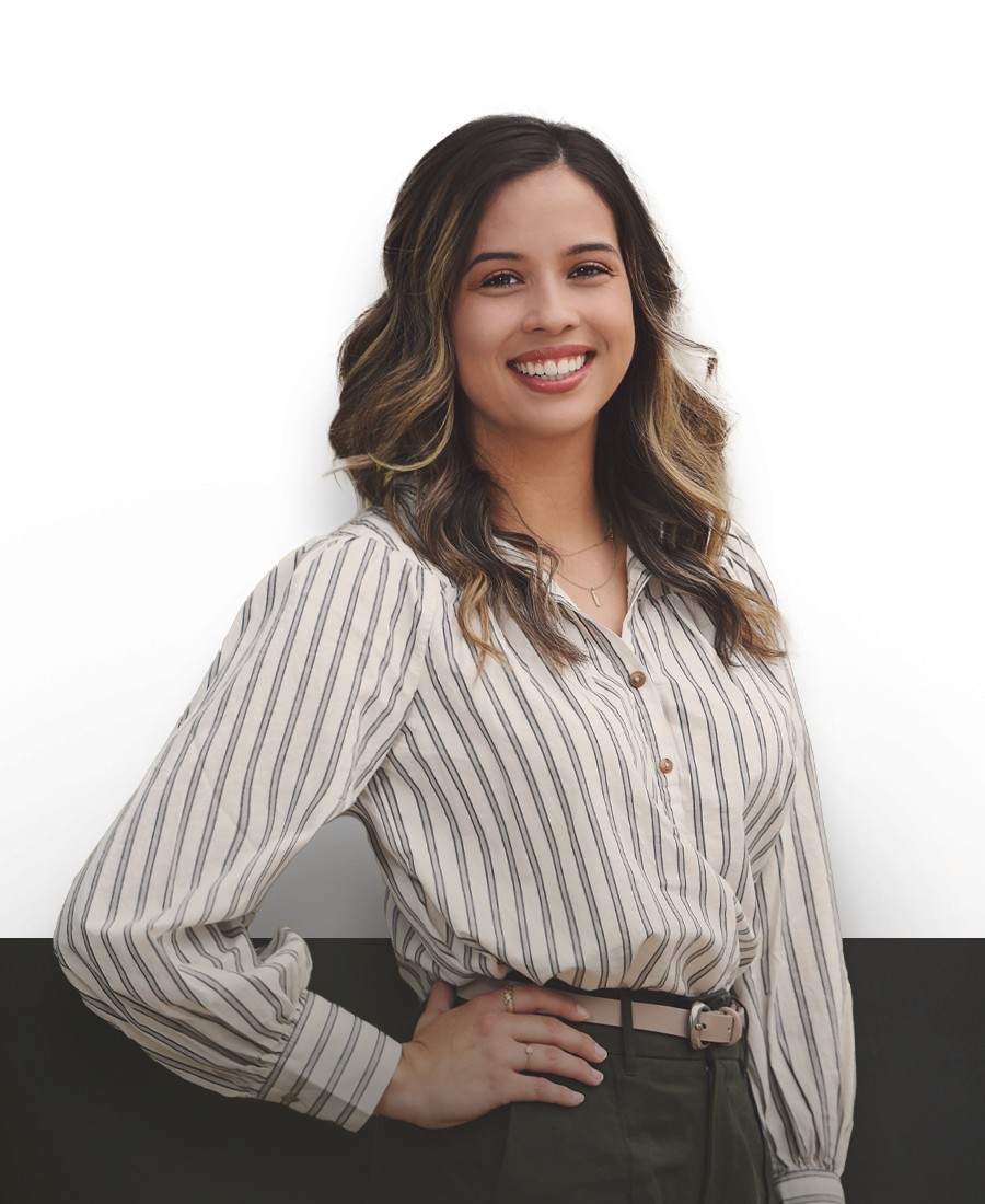 corporate headshot woman with cream and black patterned long sleeved blouse, light brown skin and brown loosely curled hair with highlights. She is posed with one hand on her hip, smiling broadly. 