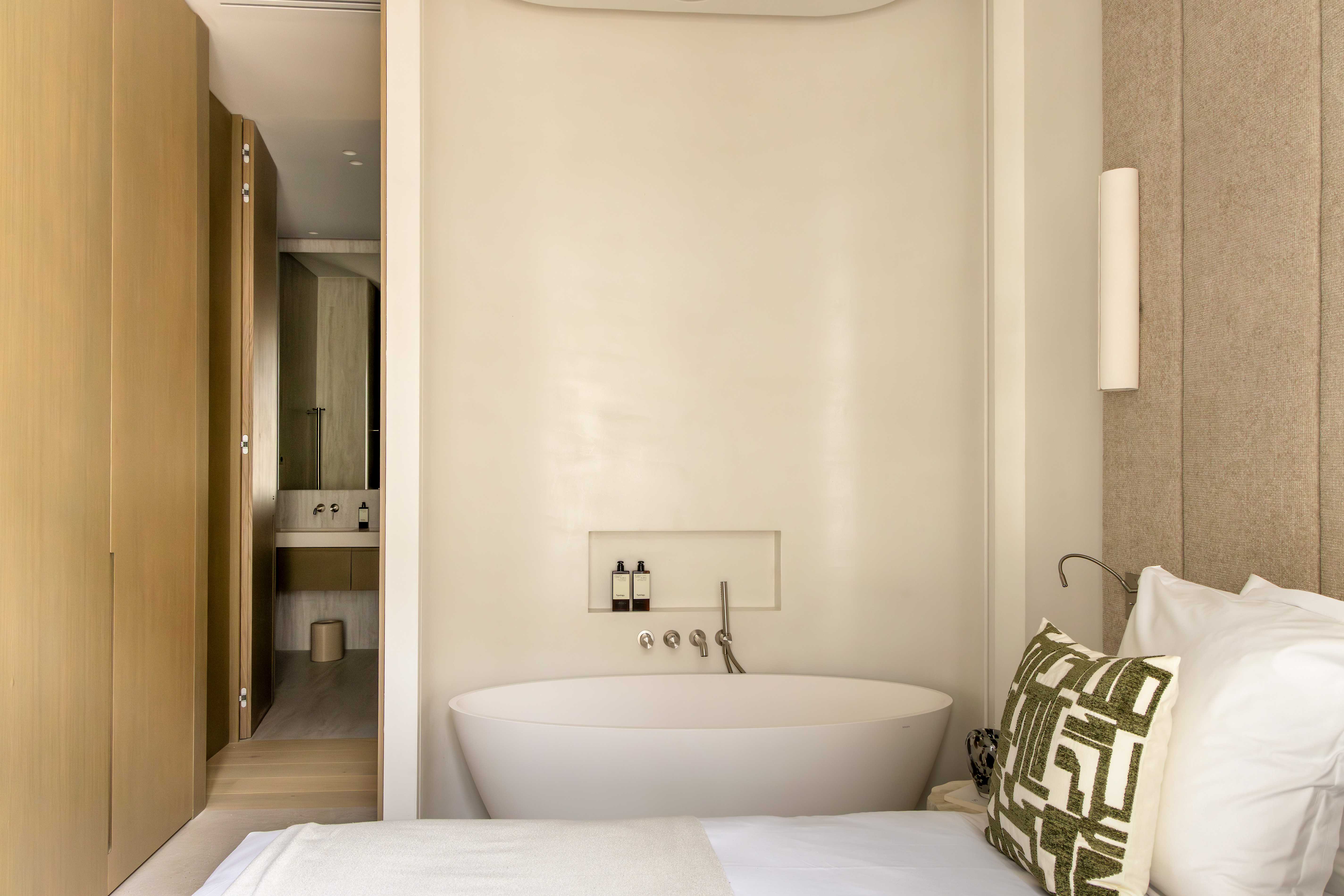 Baignoire ilot blanche dans alcove beige attenante a la chambre, avec produits de soin dans niche murale