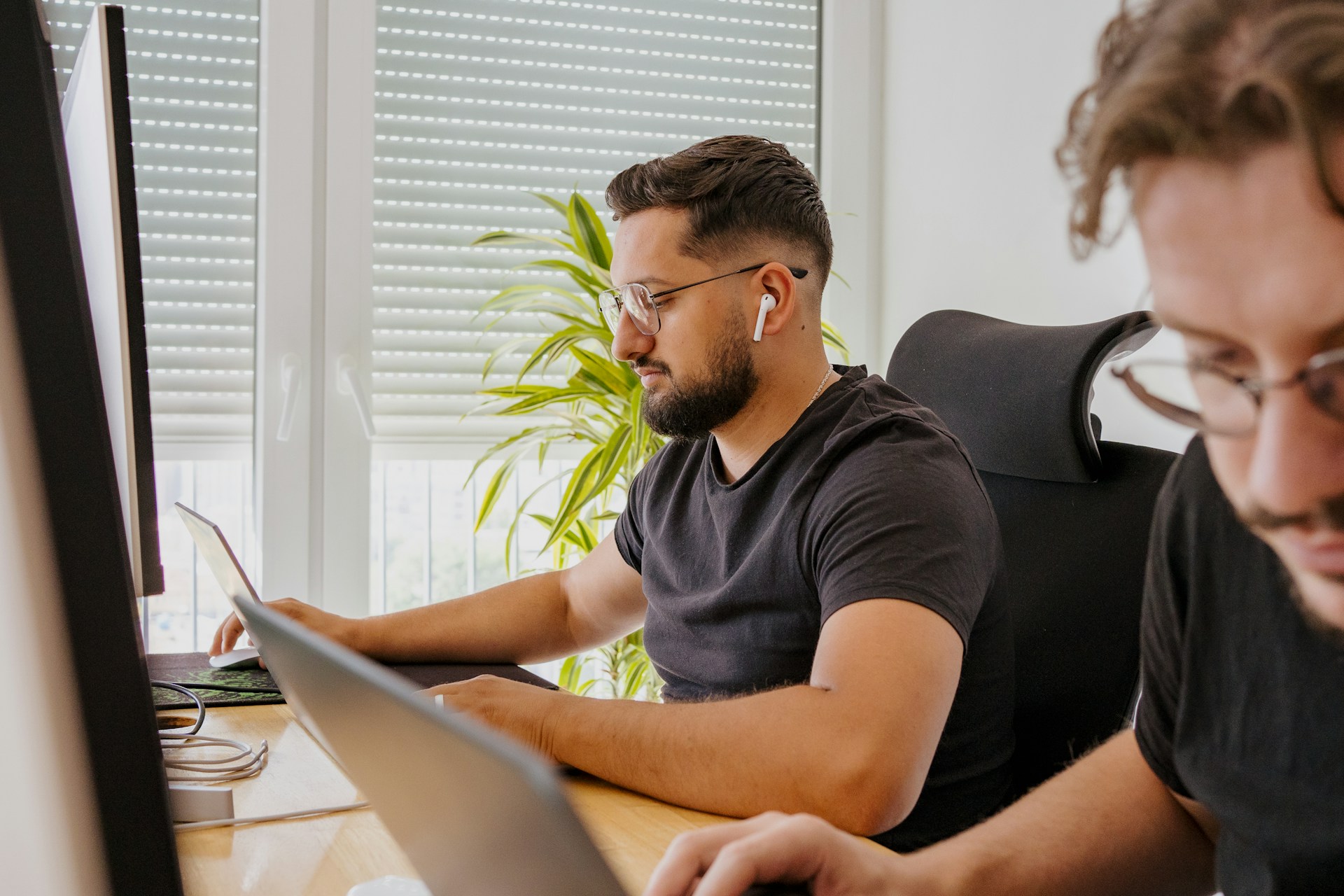 Man with glasses and an AirPod in his ear working on a laptop at a desk next to another person.