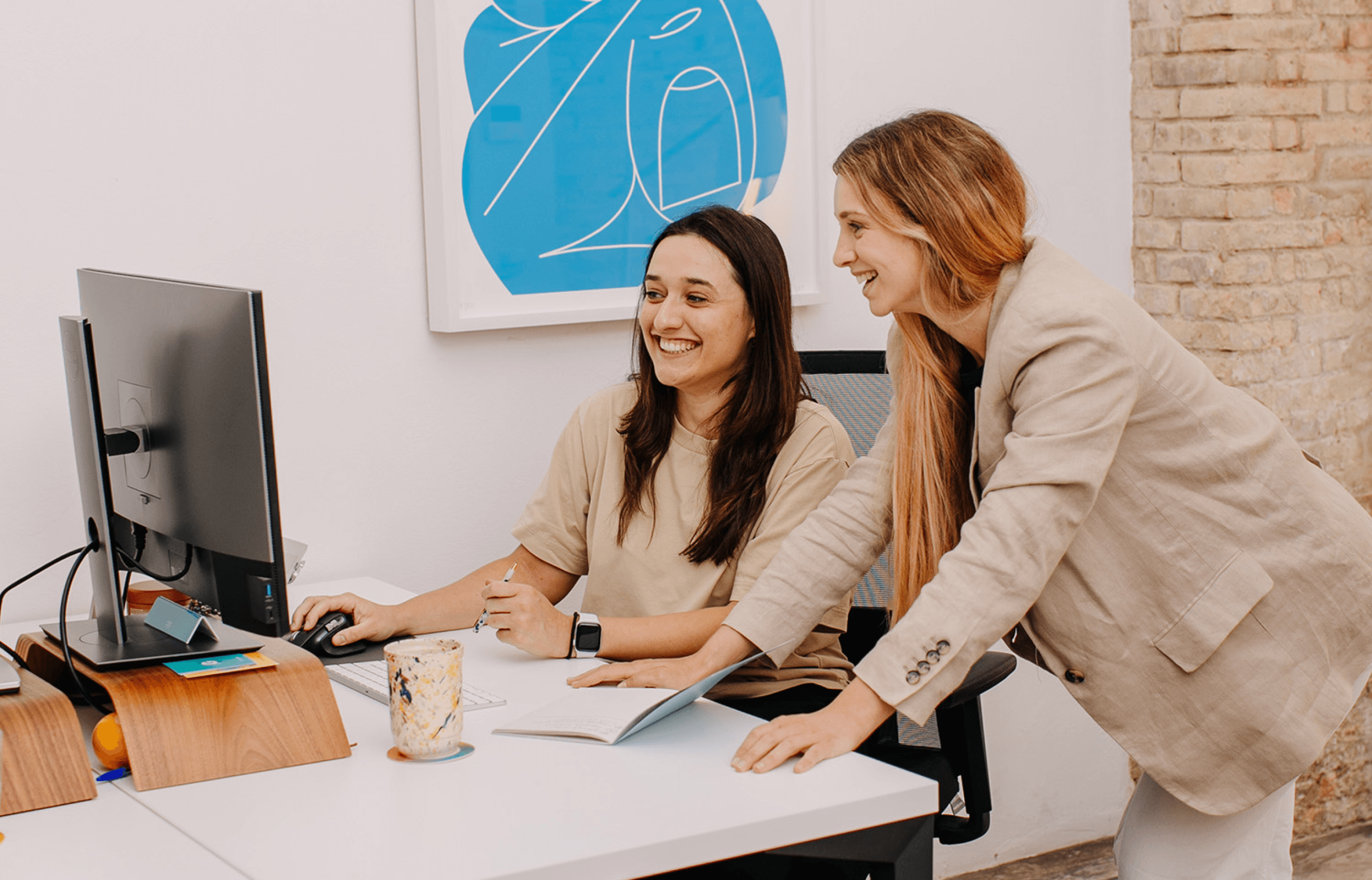 Two women looking at work on a computer, one standing and the other sitting in a chair.