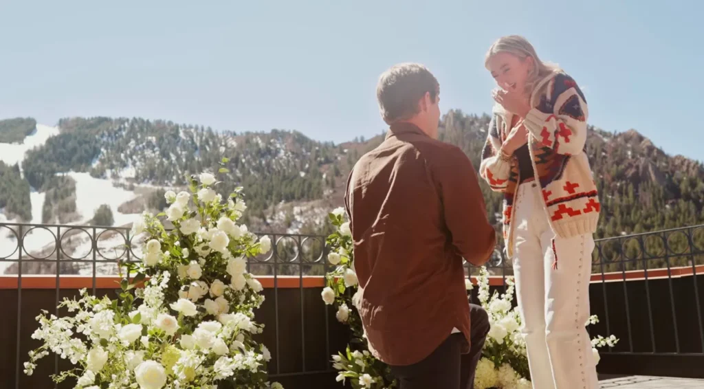 Man on one knee proposing in Snowmass Colorado on outdoor terrace with white floral arrangements and mountain views