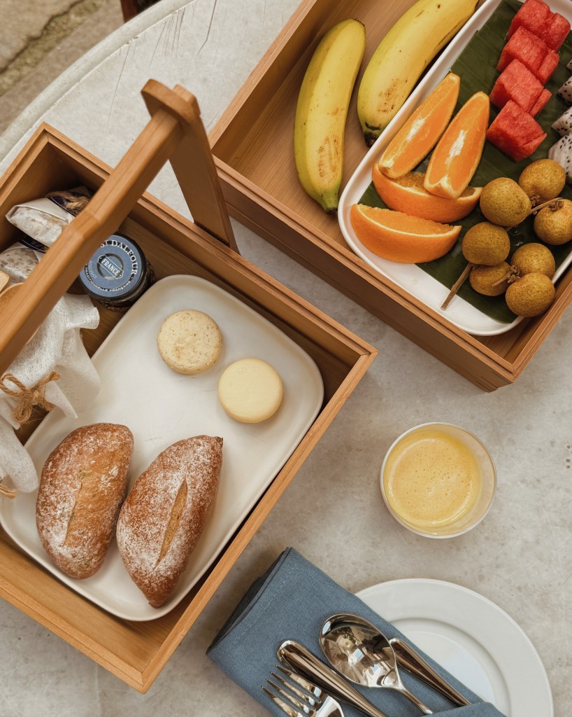 A wooden picnic box open to reveal a plate of assorted breakfast items, including two round bread rolls, a yellow butter round, and a cookie. Next to it is a separate box with bananas, orange slices, watermelon, and longan fruit arranged on a banana leaf. A glass of yellow beverage and cutlery are visible on the table.