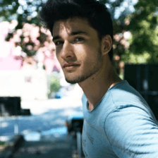 A man with dark hair and a light blue shirt stands outdoors, with sunlight filtering through tree leaves in the background, creating a serene and softly dappled effect.