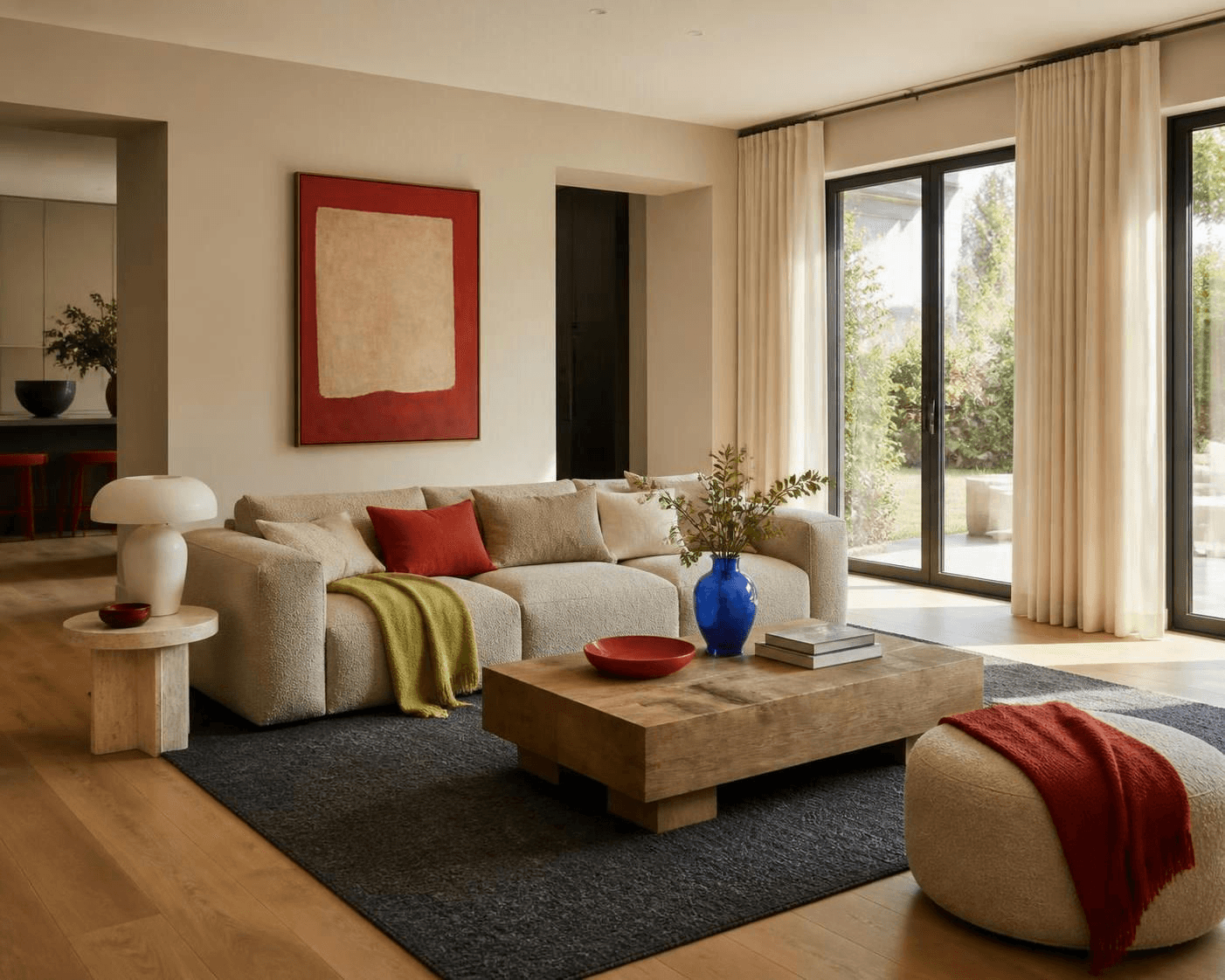 Living room with cream sofa, wood coffee table, large windows with beige curtains, red and green accents, and a blue vase with greenery.