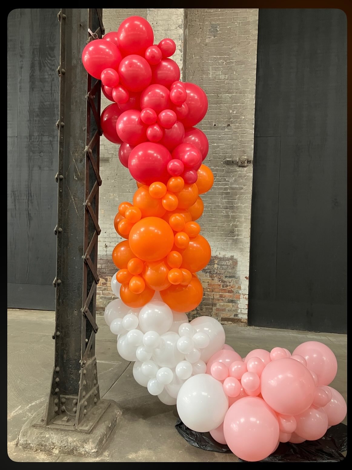 Vertical balloon garland featuring red, orange, white, and pink balloons attached to an outdoor column by Be Bold Balloons