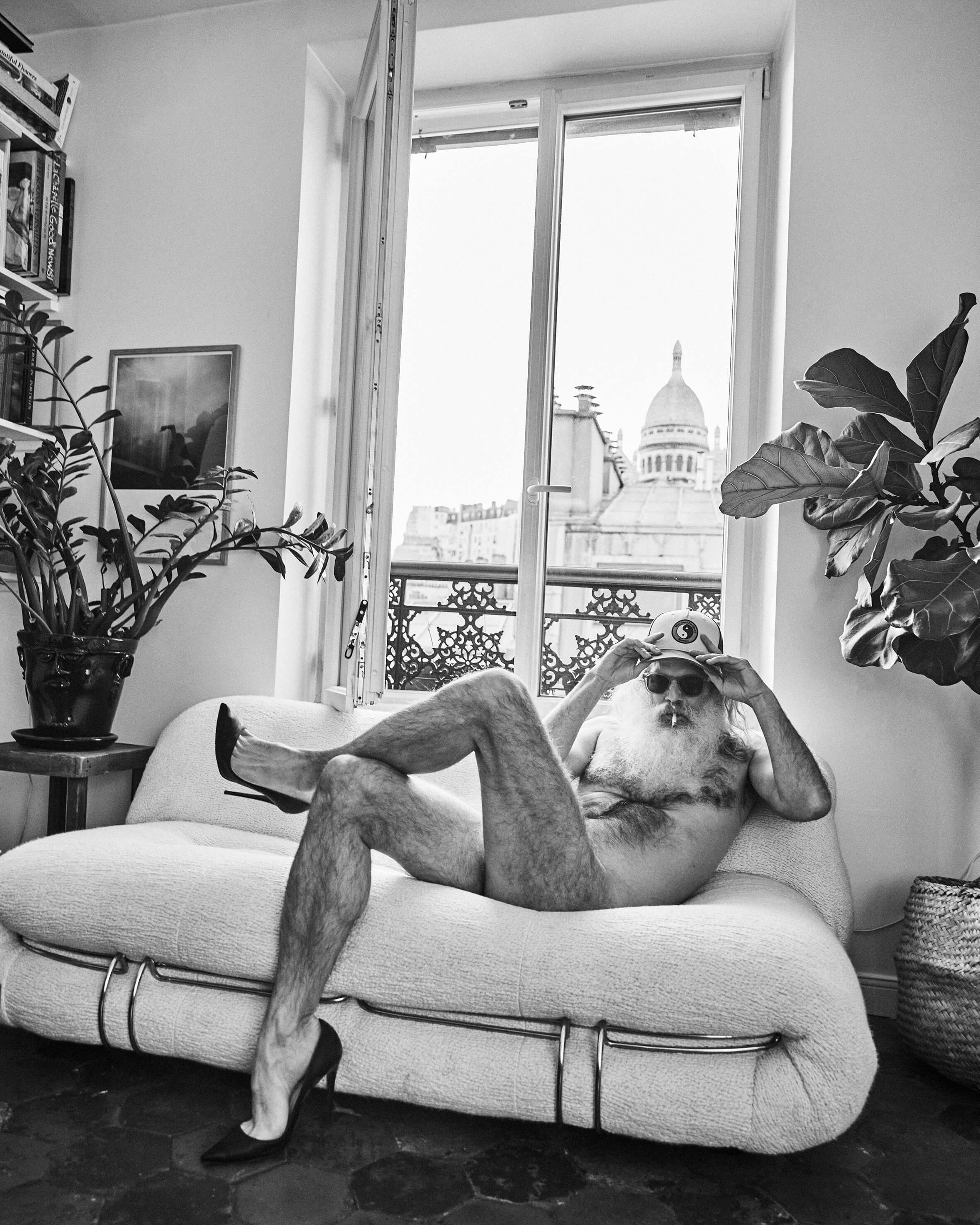 Black and white photograph by José Castellar Carayol: a nude bearded man wearing a cap and sunglasses reclines on a white Togo sofa, smoking, with the Sacré-Cœur basilica visible through an open Parisian window.