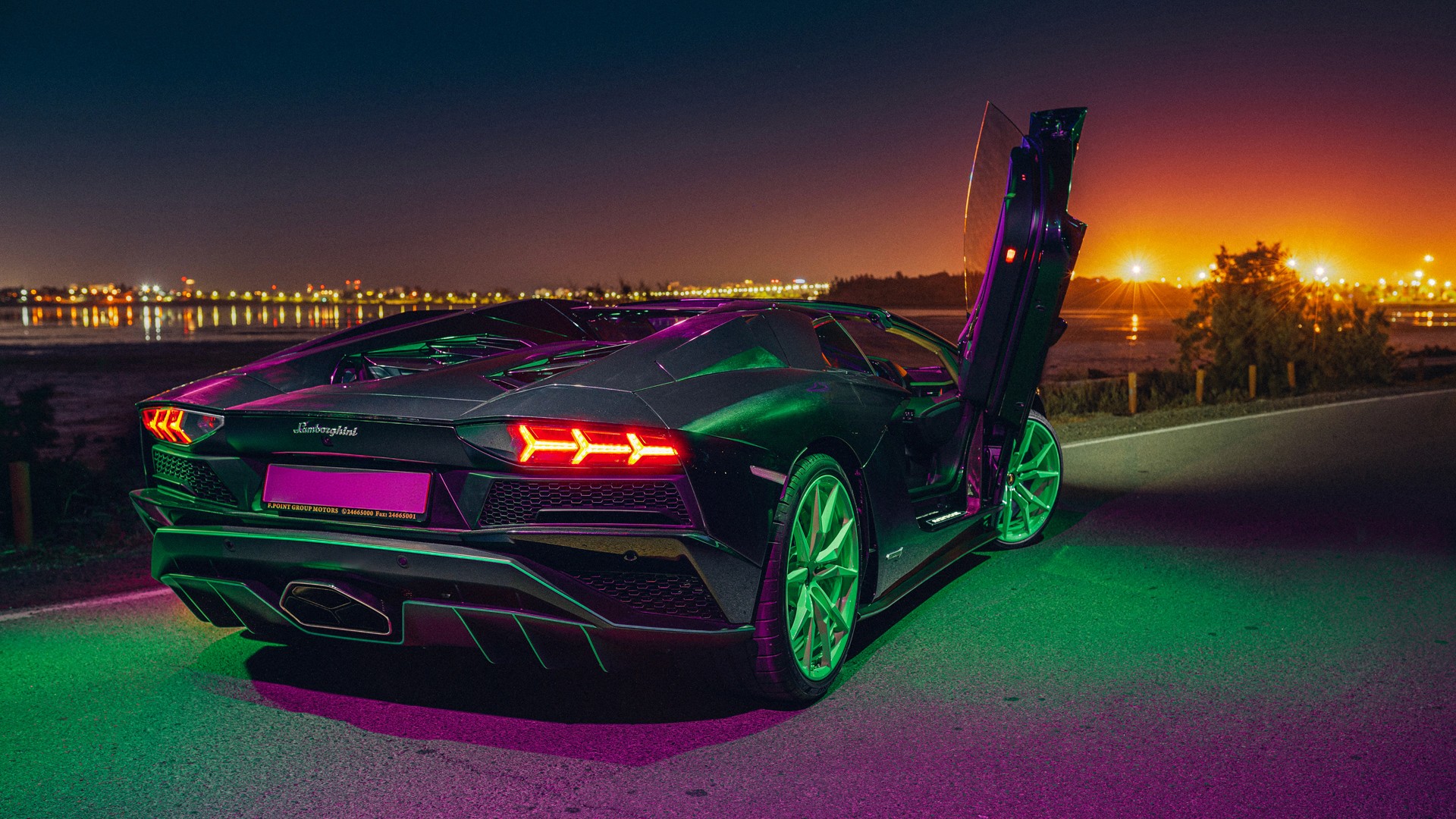 Lamborghini Aventador with green wheels and one scissor door raised, photographed from the rear three-quarter at dusk, lit with green and magenta neon, beside a waterfront with city lights reflecting across the bay and an orange afterglow on the horizon.