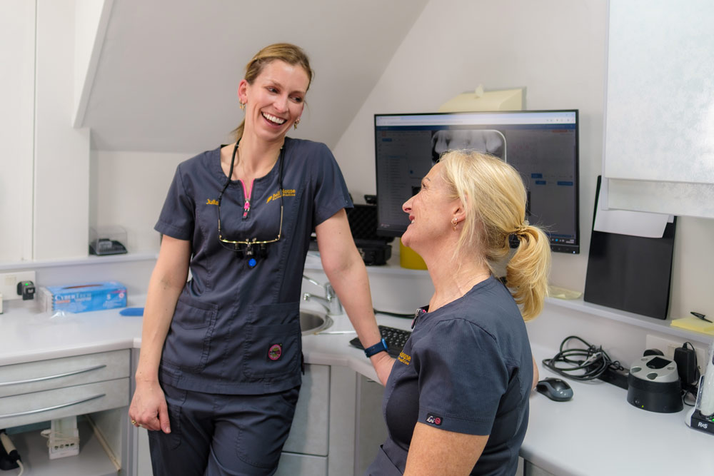 Two dental professionals, Julia and Beverley, both wearing dark blue scrubs embroidered with their names and "Bell House Dental Practice," are smiling and laughing together in a modern dental clinic. Julia, leaning against a counter, looks down at Beverley, who is sitting on a stool and looking up. In the background is a computer monitor displaying a dental x-ray, a sink, and various dental equipment.