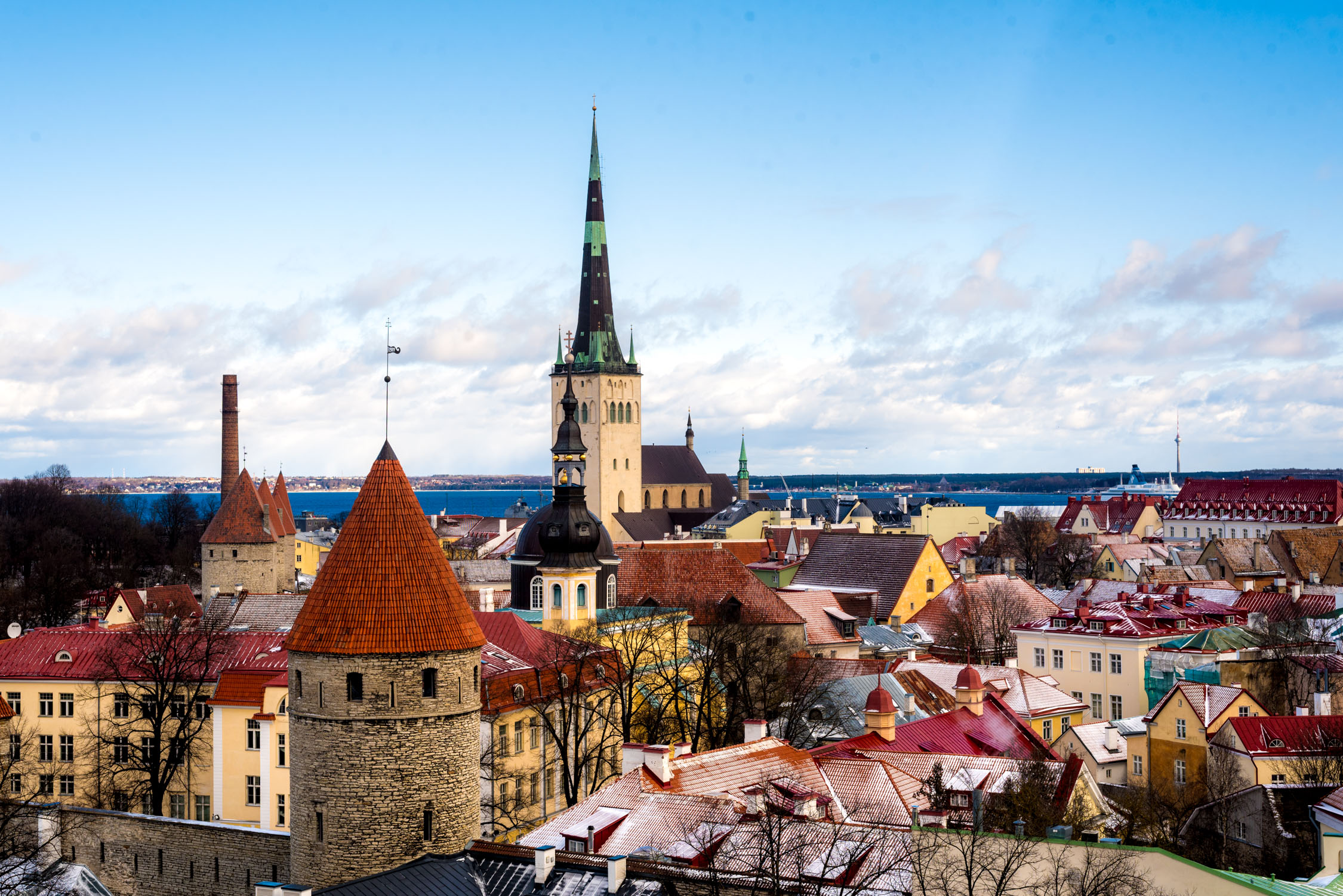 Colorful rooftops and tall spires rise against a bright, partly cloudy sky over a densely built historical settlement.