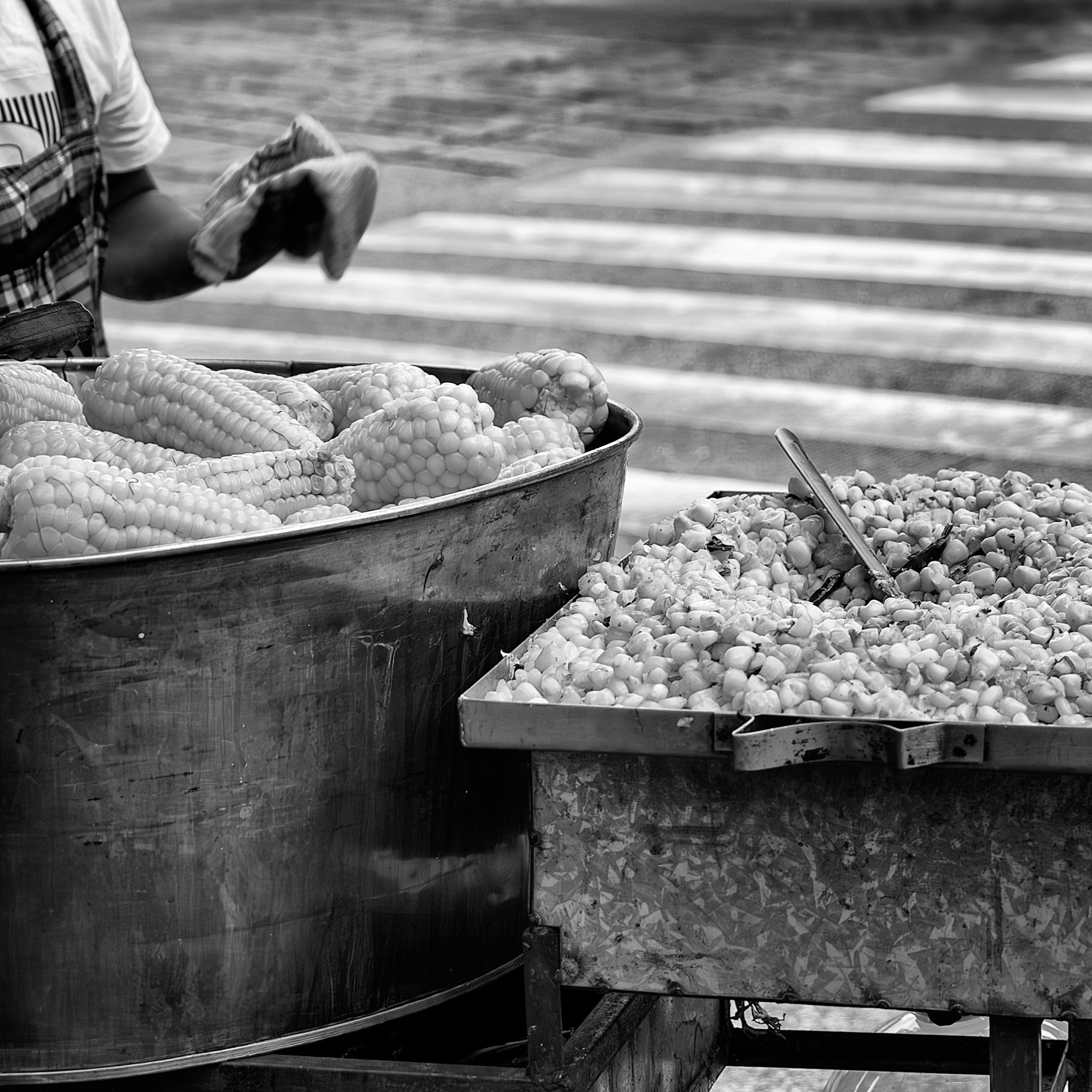 Black and white photo of a street vendor's cart with a large pot of steamed corn on the cob and a metal tray of corn kernels ready to be served, set near a crosswalk.