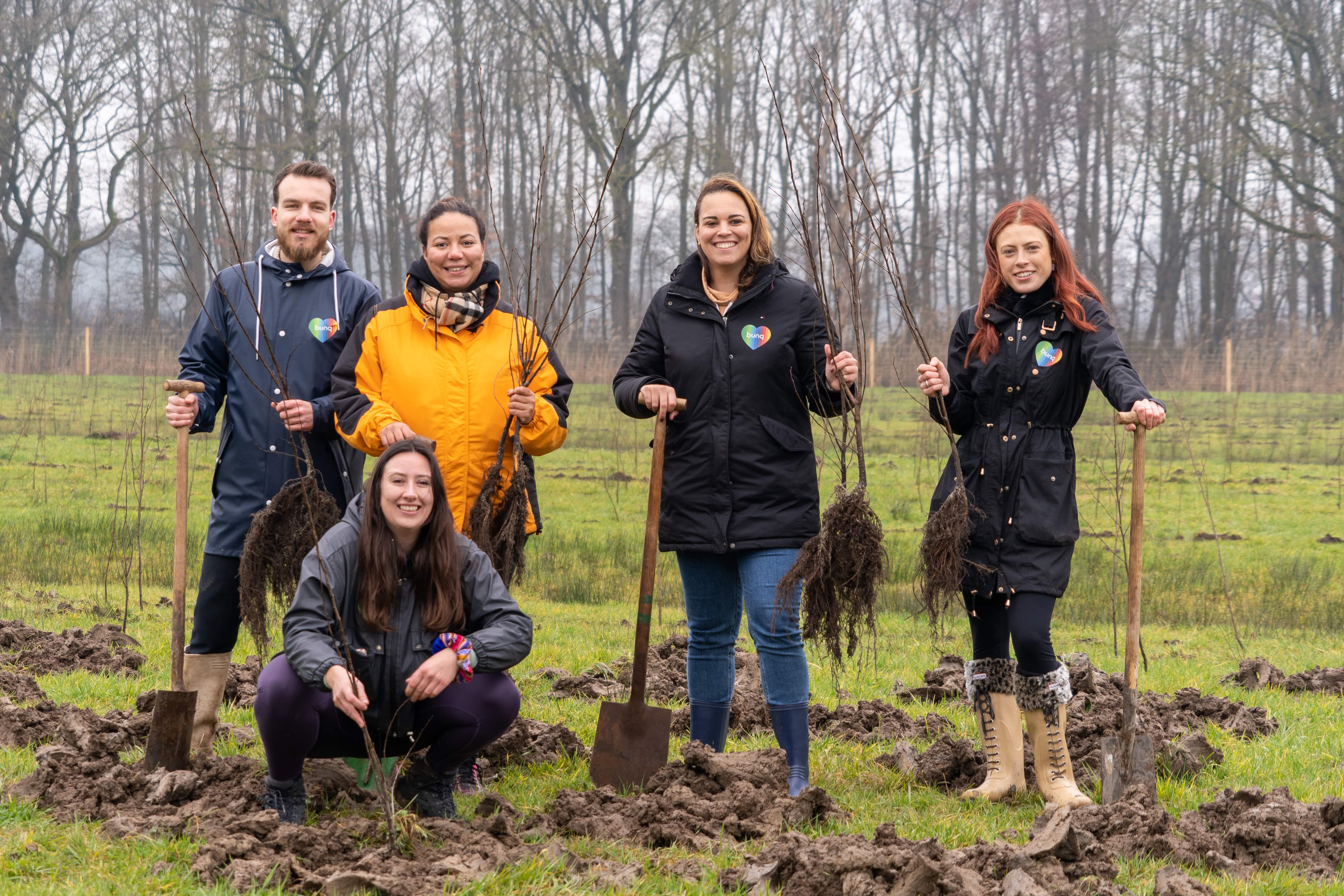 Een groep van vijf mensen in werkkleding staat op een modderig veld, met gereedschap in hun handen en glimlachend naar de camera.