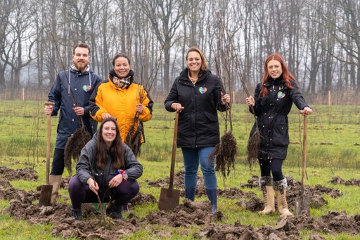 A group of five people in work clothing stands on a muddy field, holding tools and smiling at the camera.