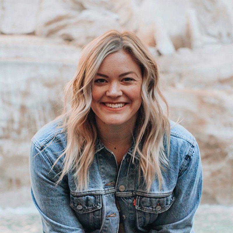 A smiling woman with blonde, wavy hair wearing a denim jacket, positioned in front of a blurred stone fountain or rocky background.
