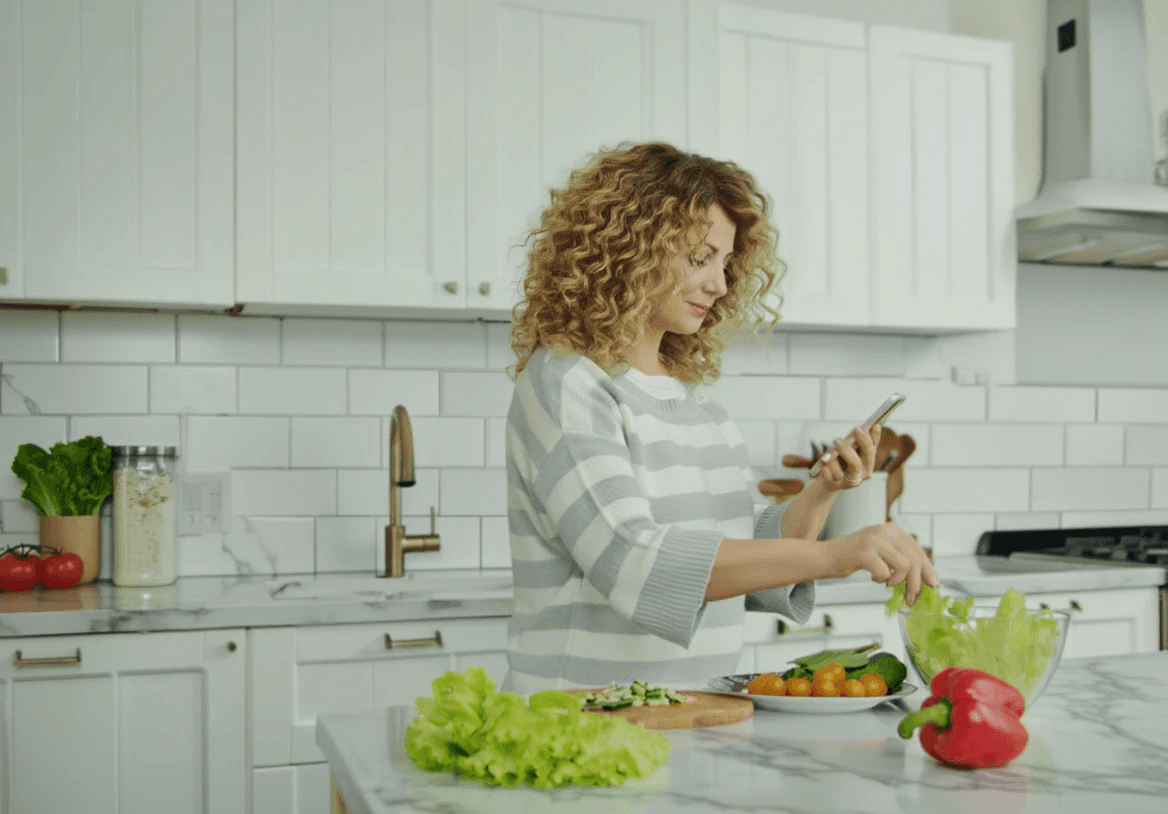 En kvinna står i sitt kök och förbereder hälsosam, näringsrik mat medan hon tittar på sin mobil