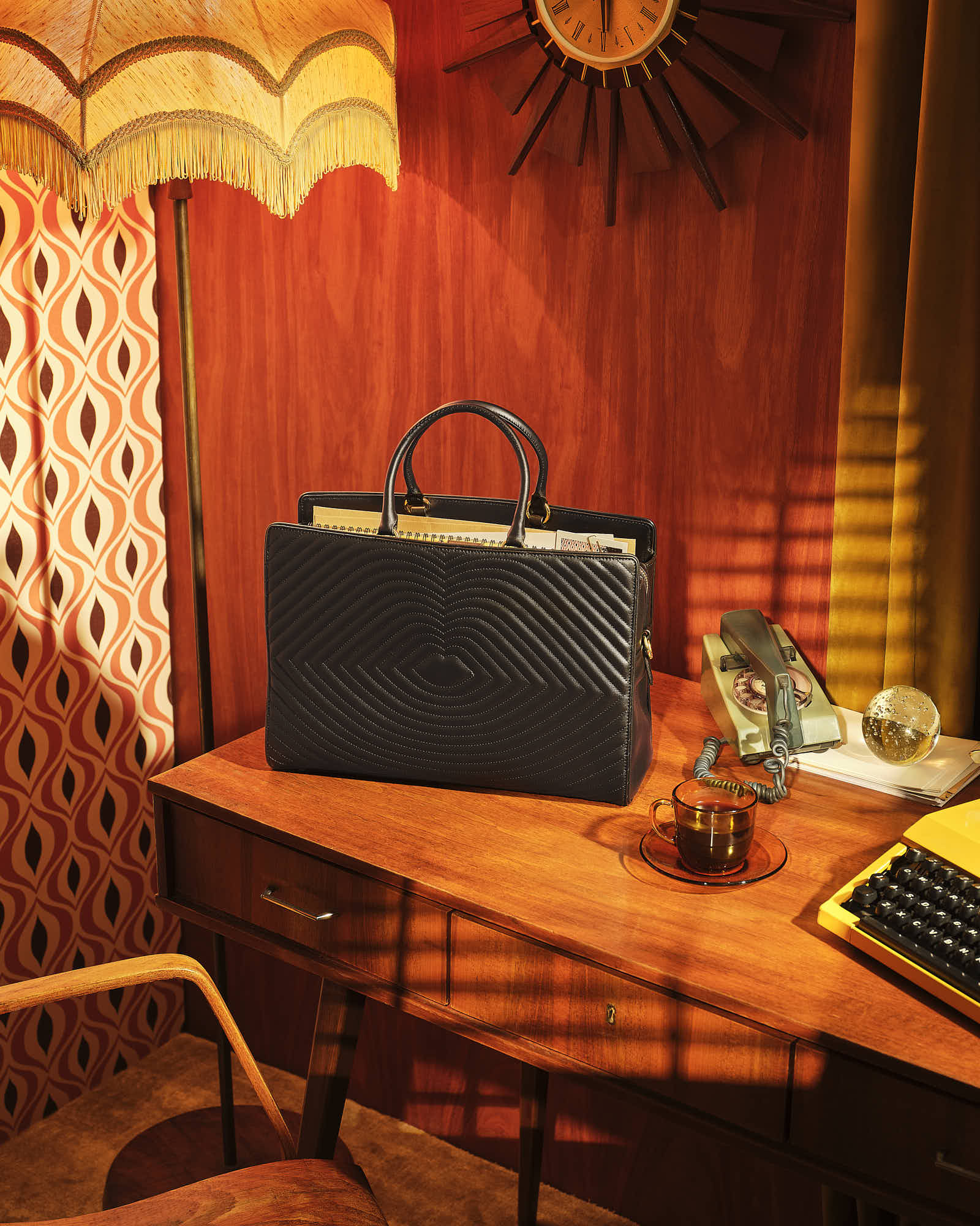 Vintage office setting with a stylish black handbag on a wooden desk featuring a rotary phone, tea cup, typewriter, and mid-century decor.