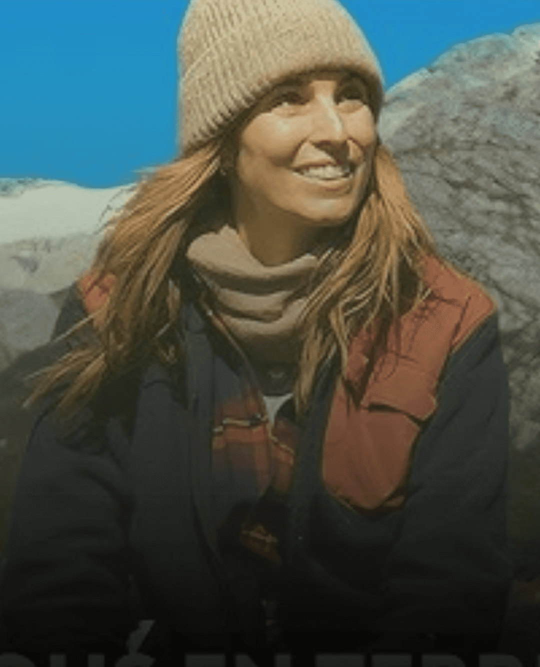 A smiling woman with curly hair is posing in a warmly lit indoor setting. She wears a dark-colored shirt.