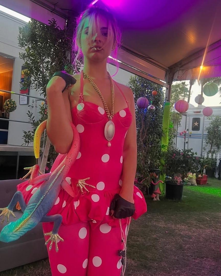a person wearing a bright pink polka dot outfit holds a colorful lizard prop