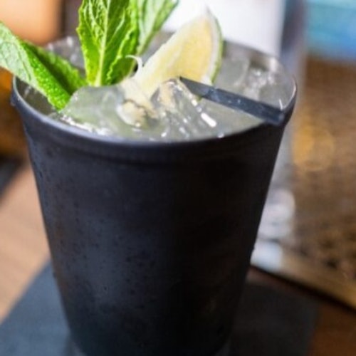 Una taza negra con una ramita de menta, una rodaja de limón y un popote en una bebida clara, sobre una servilleta en una superficie de madera.