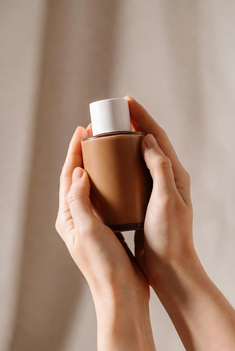 Two hands holding a glass bottle of tan liquid foundation with a white cap against a neutral backdrop.