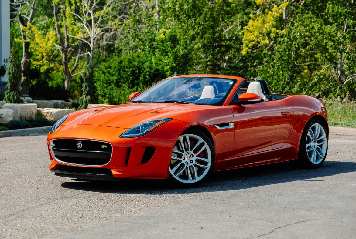 Une Jaguar F-Type cabriolet orange posée en plein soleil, avec en arrière-plan une végétation dense. La silhouette fluide et les lignes tendues du roadster mettent en avant son caractère sportif, élégant et expressif.